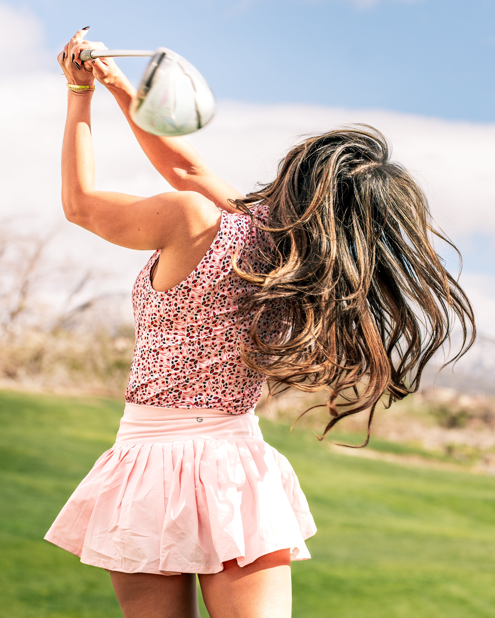 Woman swinging a golf club in a floral tank top and pink skirt, showcasing a stylish and confident golf look.