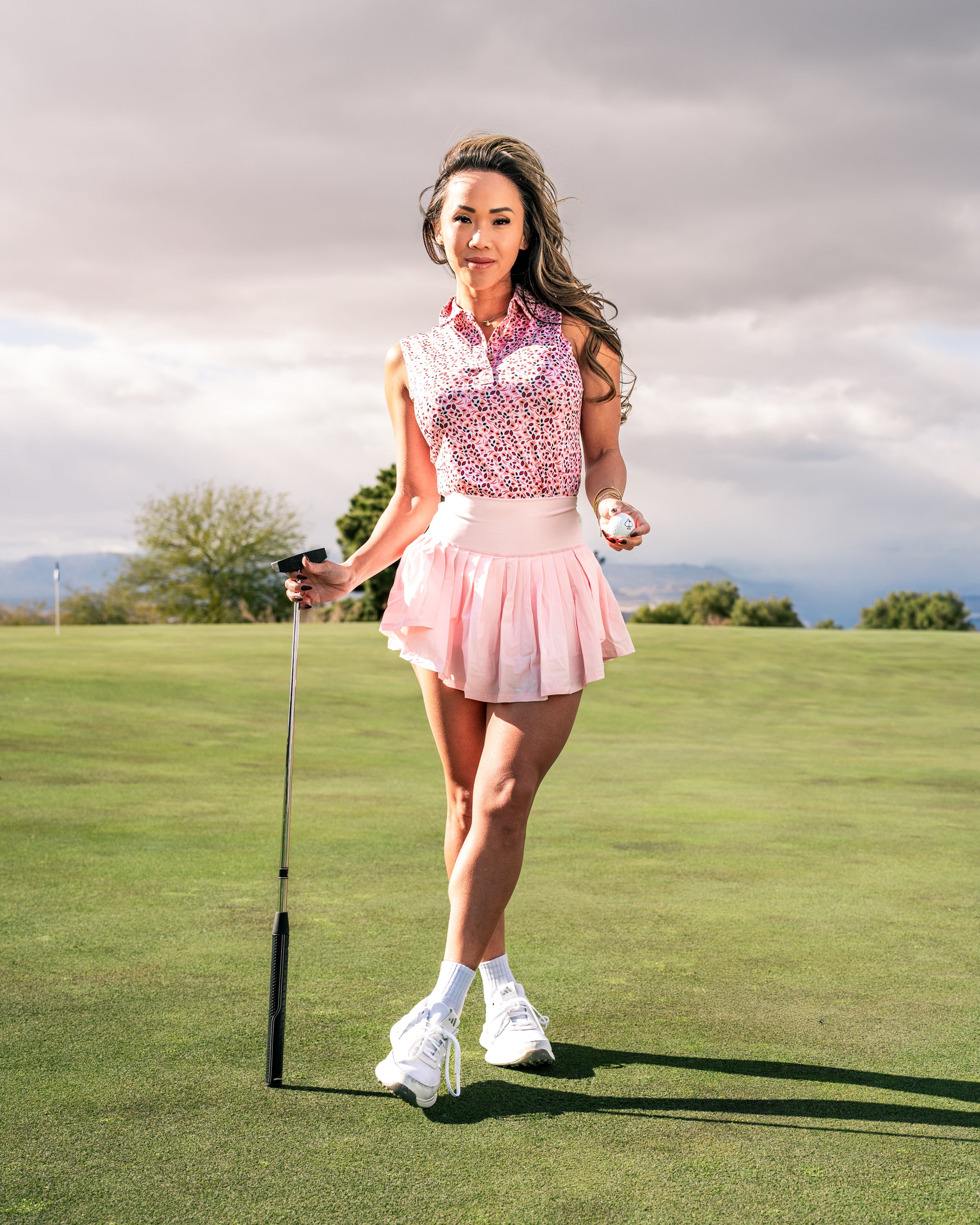 A woman stands on a golf course wearing a pink floral golf polo and a matching skirt, holding a golf ball and club.