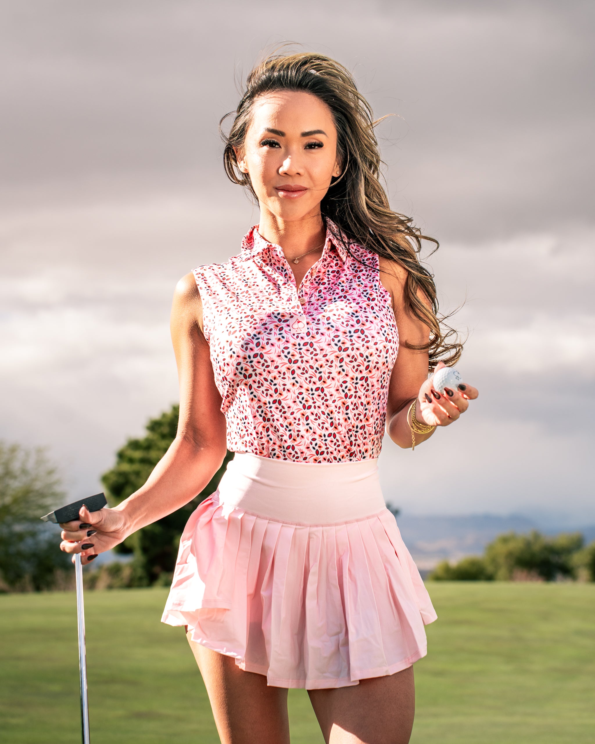 Stylish woman in a floral golf polo and pink skirt, holding a putter on a golf course under cloudy skies.