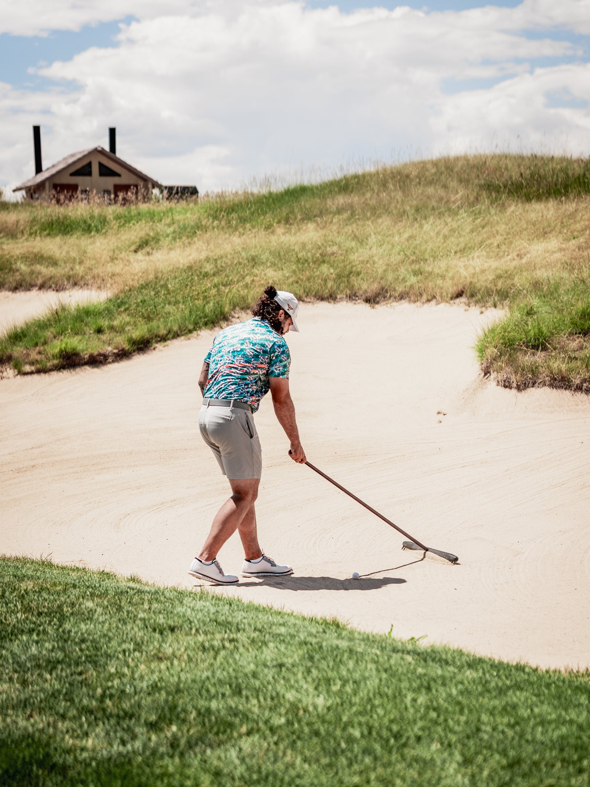 A golfer in a colorful Trout Splatter shirt rakes sand in a bunker, showcasing bold golf apparel on the course.