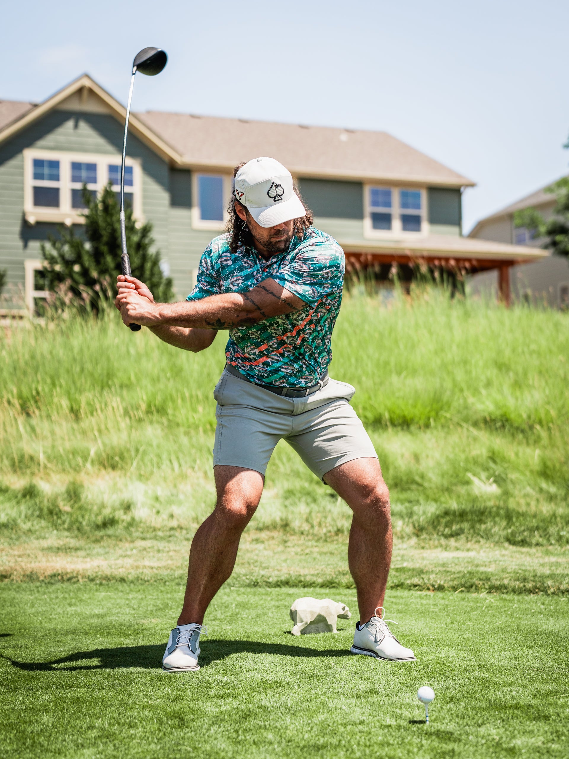 A golfer in a vibrant Trout Splatter shirt and cap prepares to swing a driver on a sunny course.