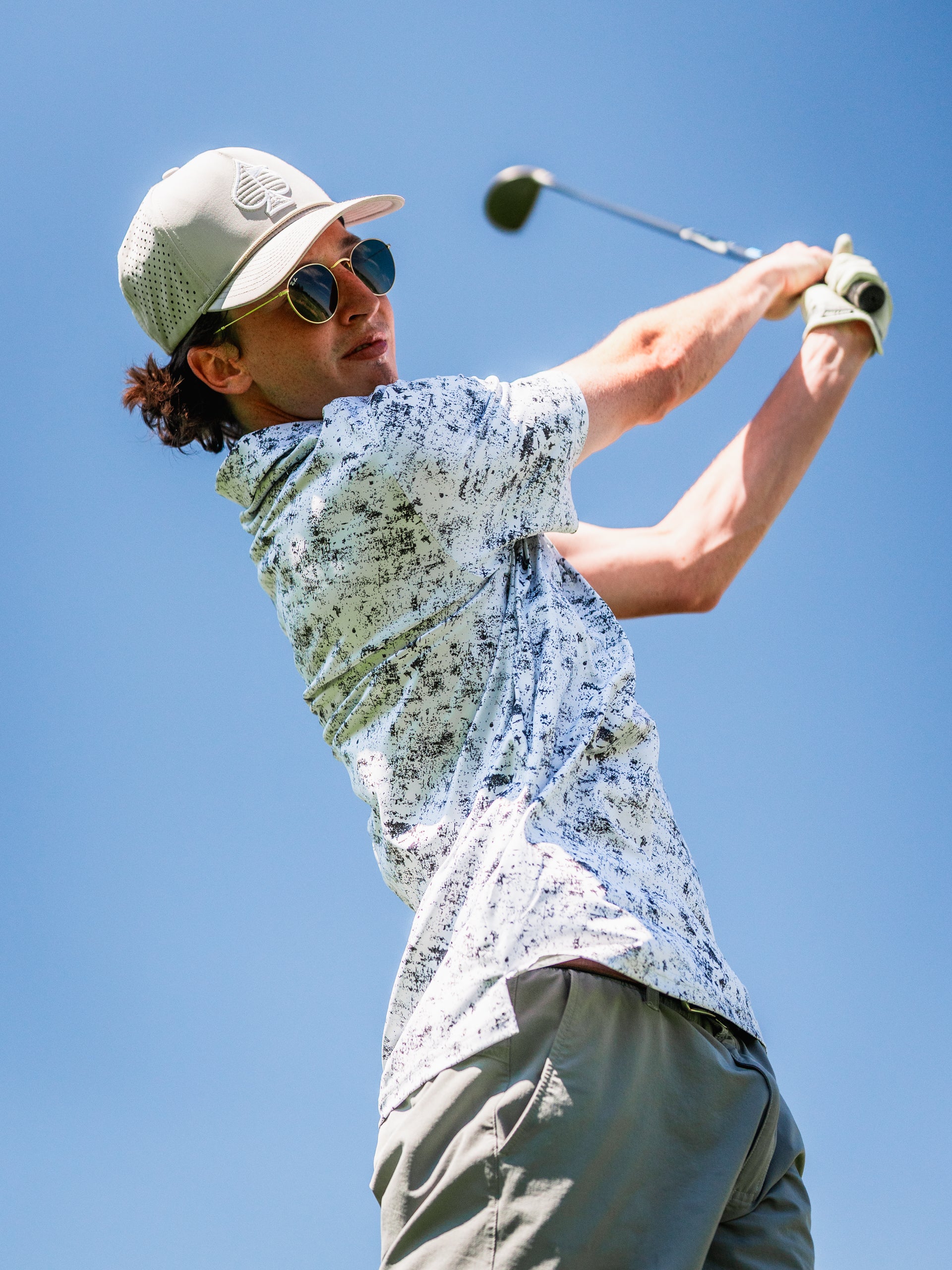 A golfer swings a club on a sunny day, wearing a stylish patterned shirt and a cap, showcasing a bold golf look.