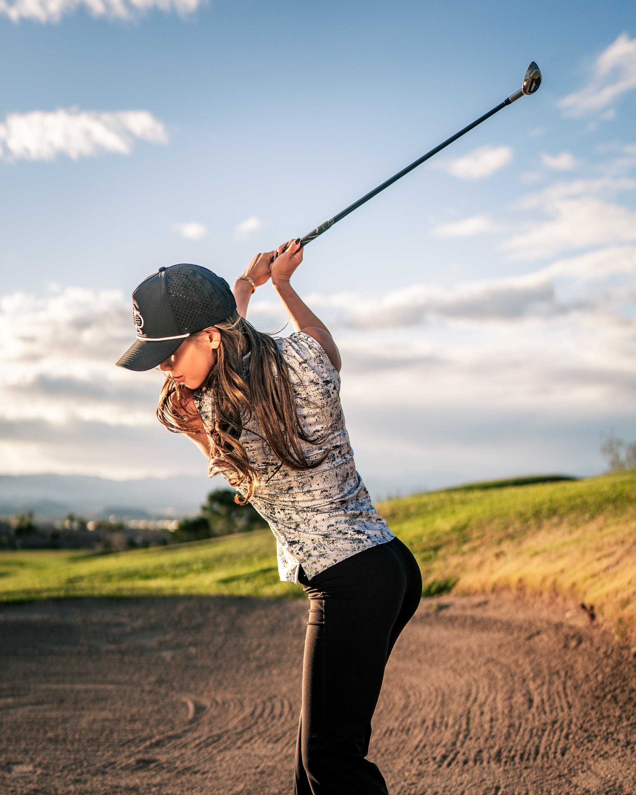 A woman swings a golf club on a course, wearing a stylish patterned golf shirt and cap, showcasing bold golf apparel.