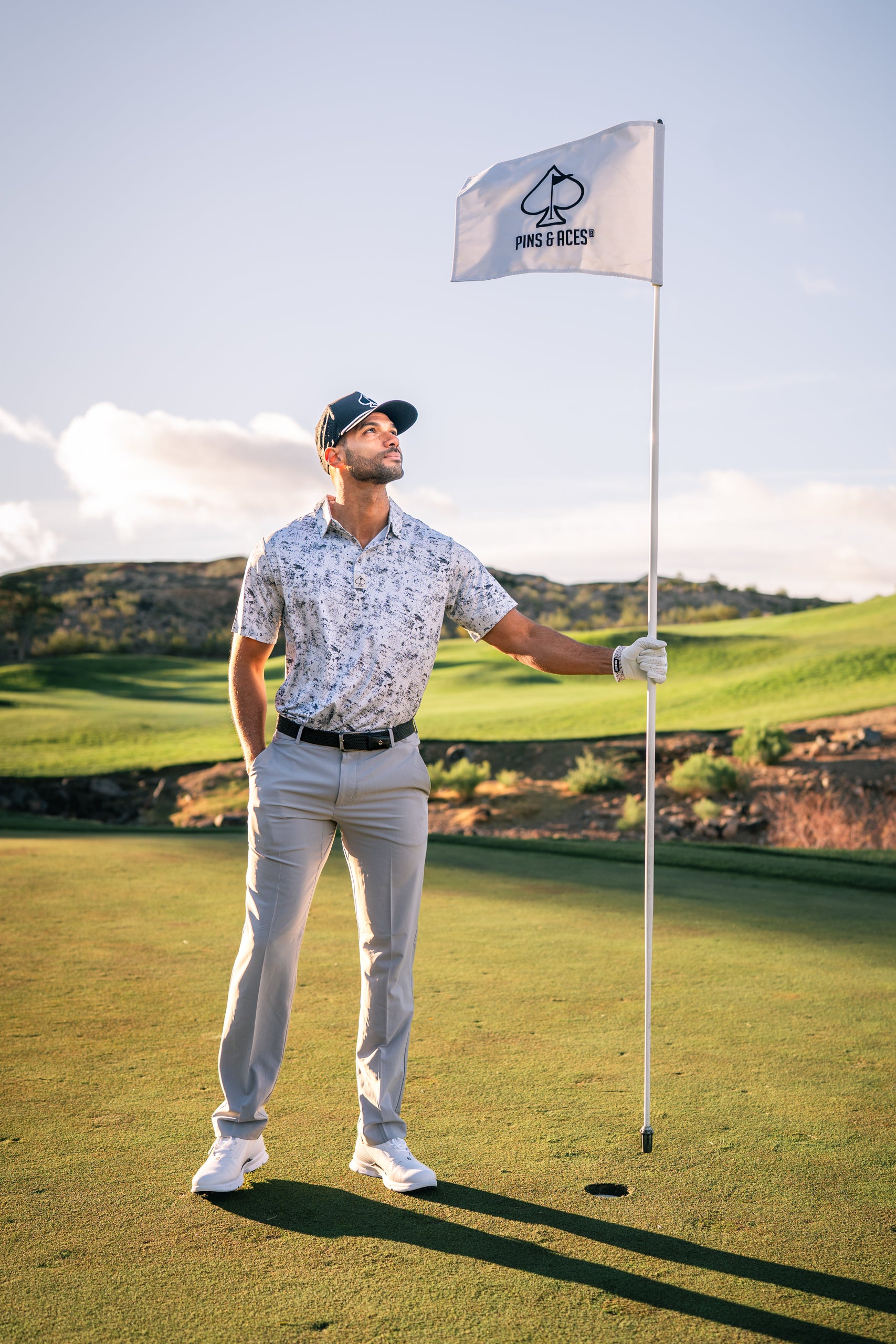 A golfer in a stylish white floral polo shirt and light gray pants stands confidently by a golf flag on the course.