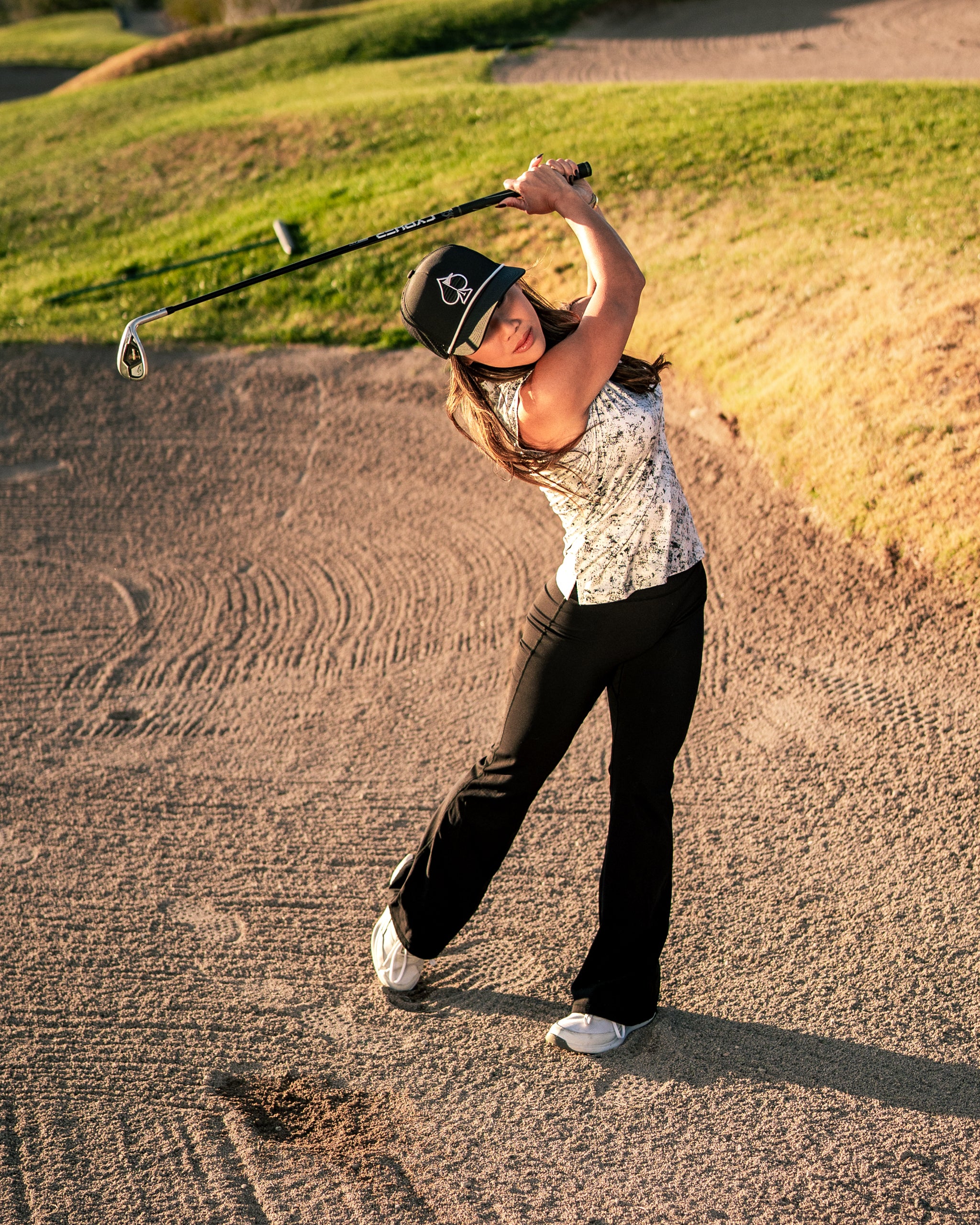 A woman swings a golf club on a course, wearing a stylish patterned top and black pants, showcasing bold golf apparel.