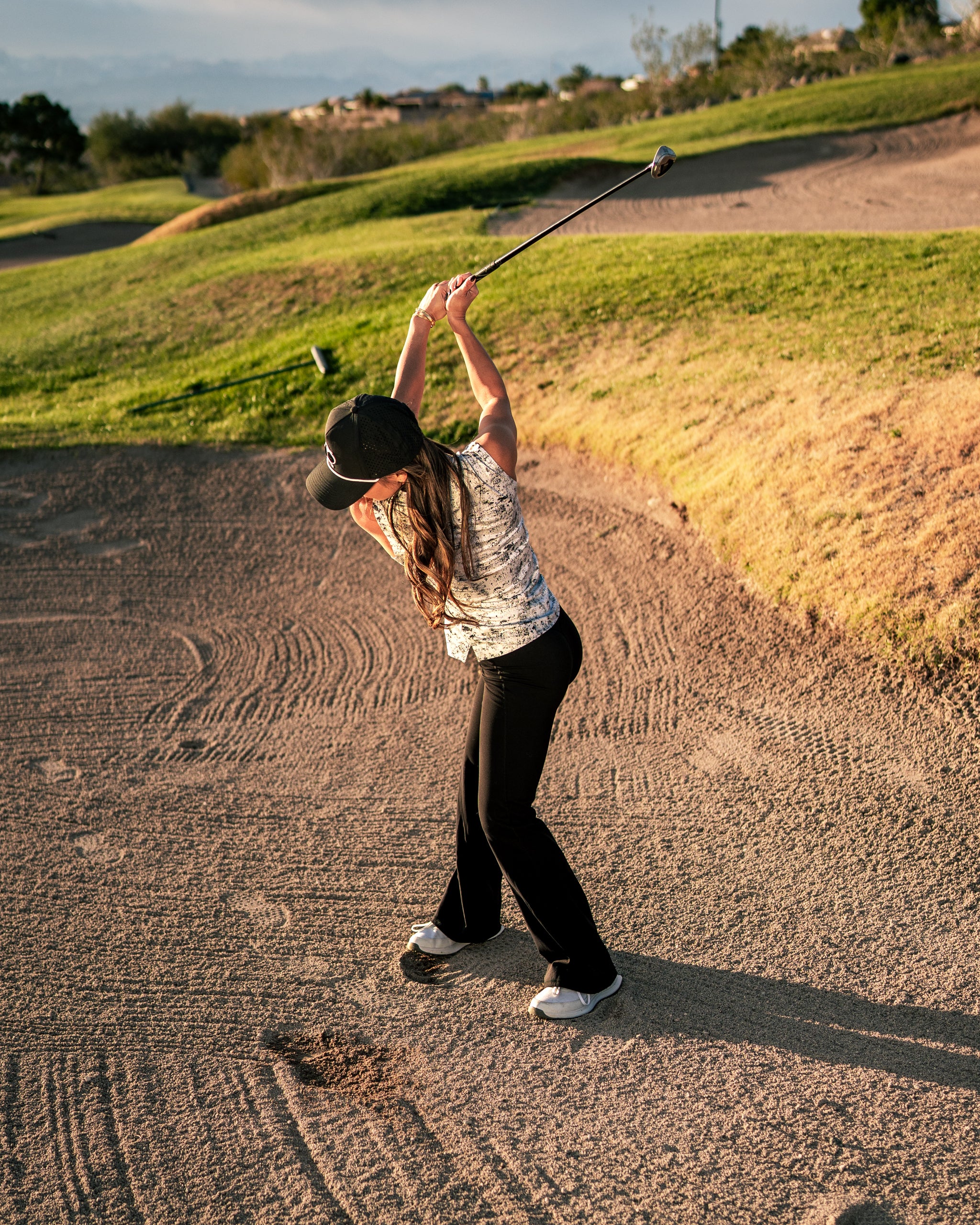 A woman swings a golf club in a sand trap, wearing a stylish patterned top and black pants, showcasing bold golf apparel.