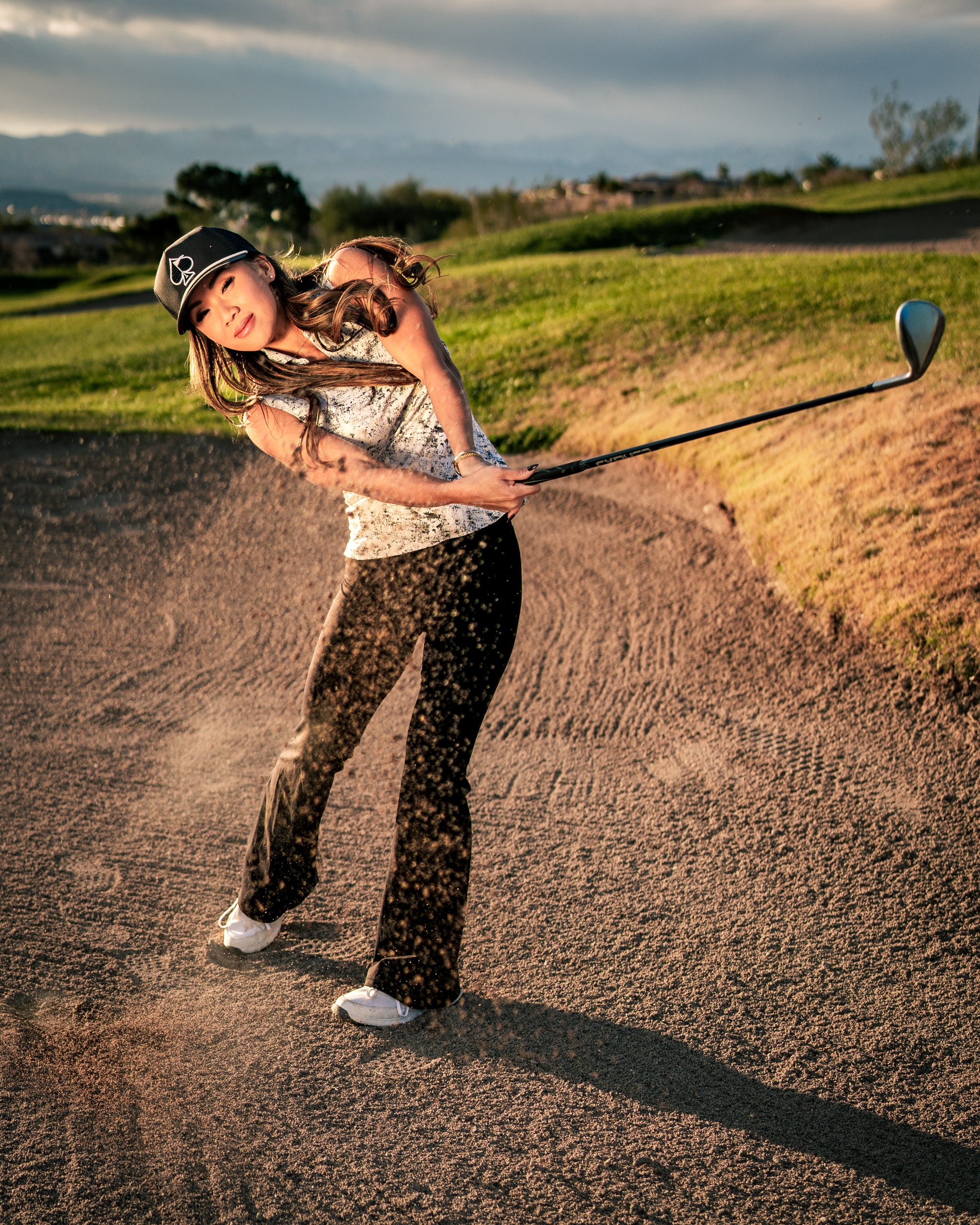 A woman in a stylish golf outfit swings a club from a sand trap, showcasing confidence and skill on the course.