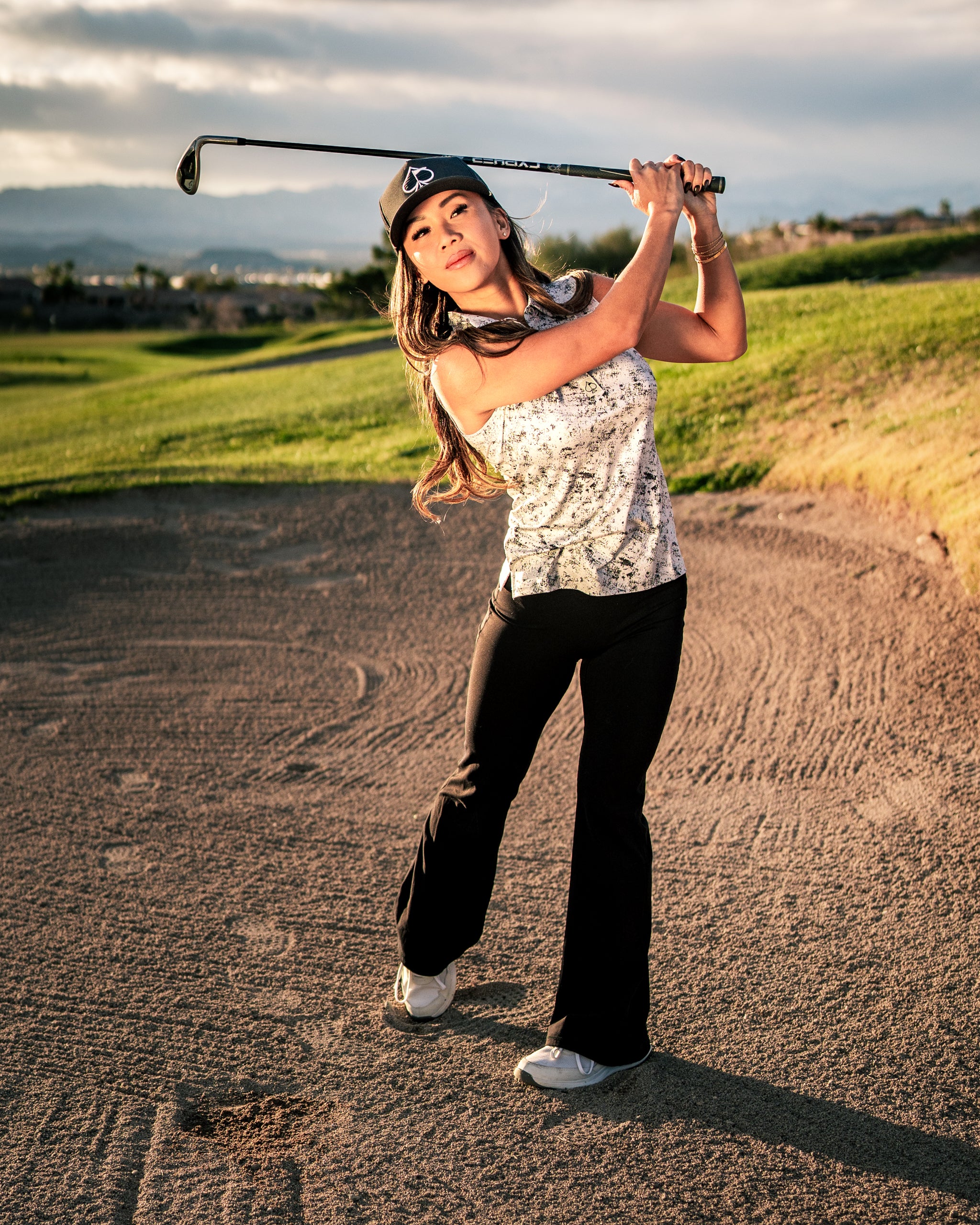 Woman in a stylish golf outfit swings a club on a sandy course, showcasing the Women's White Noise apparel.