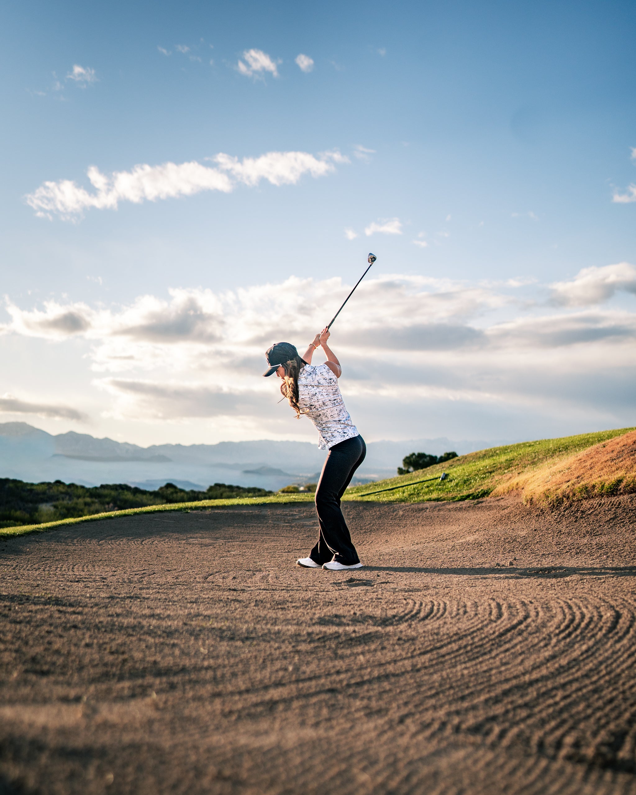 A woman swings a golf club on a sandy course, wearing a stylish white golf shirt and black pants against a scenic backdrop.