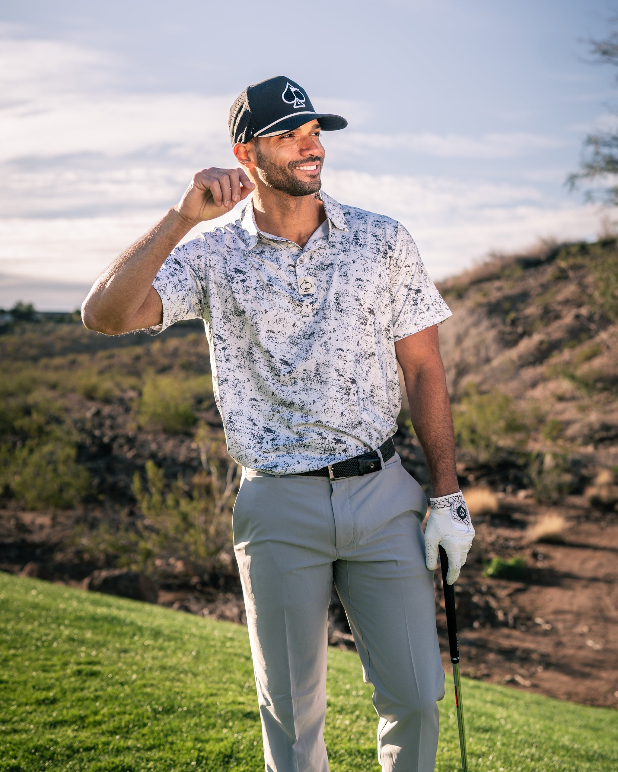 A golfer smiles while wearing a white and gray patterned polo shirt and light-colored pants on a sunny course.