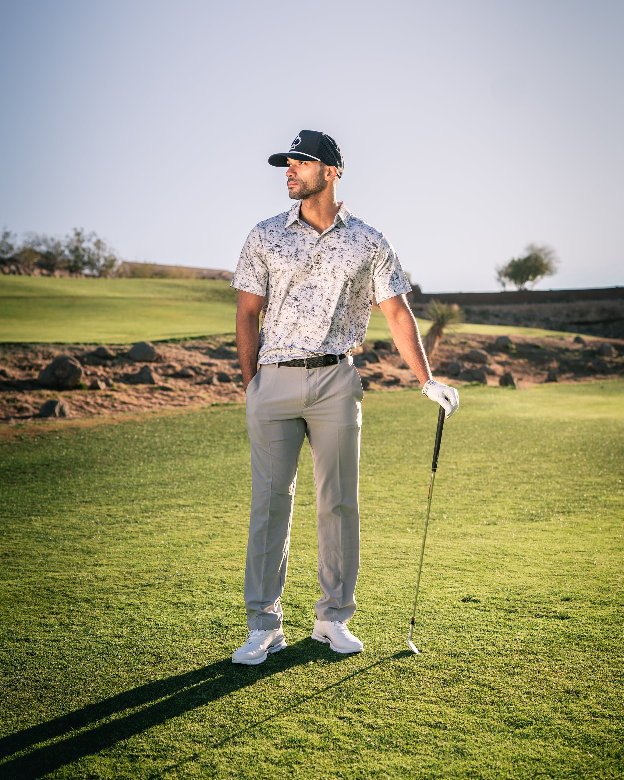 A golfer stands confidently on the course, wearing a stylish white noise patterned shirt and light-colored pants.