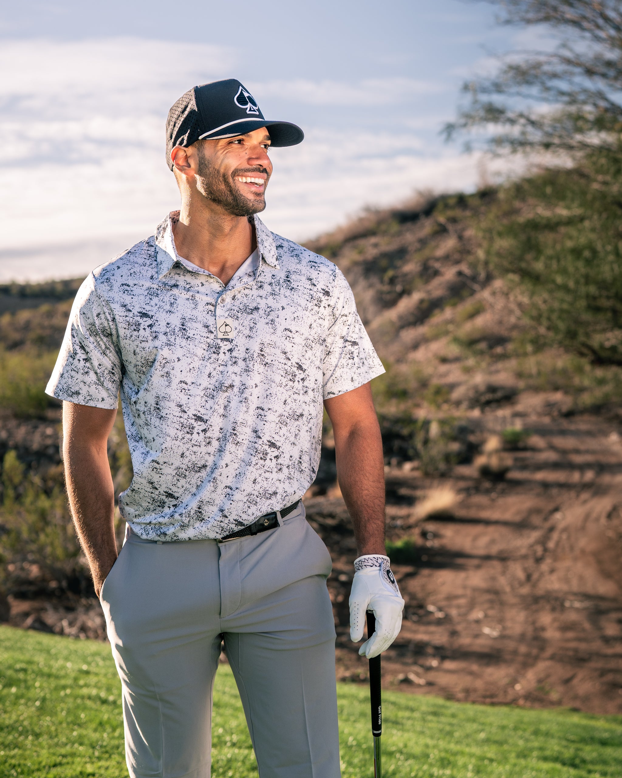 A golfer smiles while wearing a stylish white noise patterned polo shirt and gray pants on a sunny course.