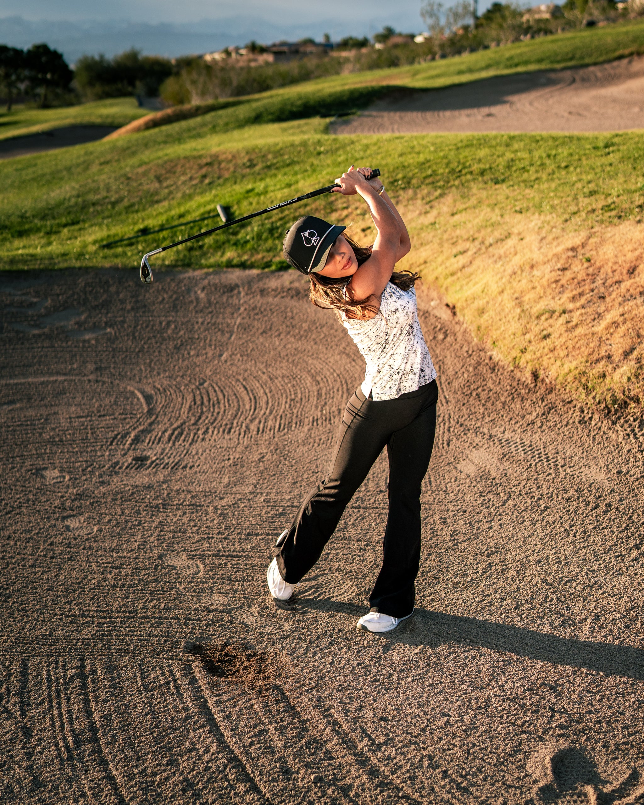 A woman swings a golf club on a course, wearing a stylish white top and black pants, showcasing bold golf fashion.