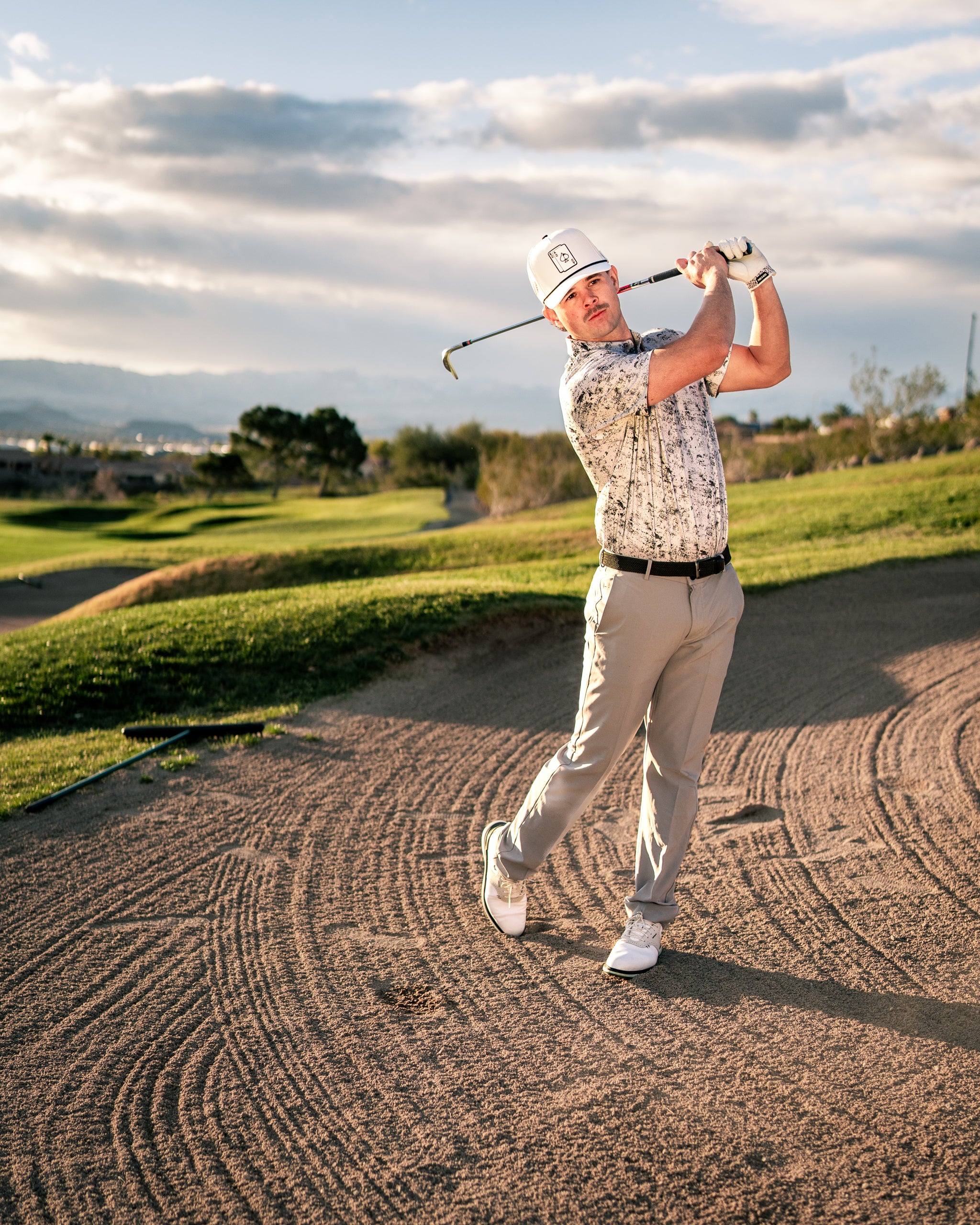 A golfer swings a club on a sandy course, wearing a stylish white patterned shirt and light gray pants.