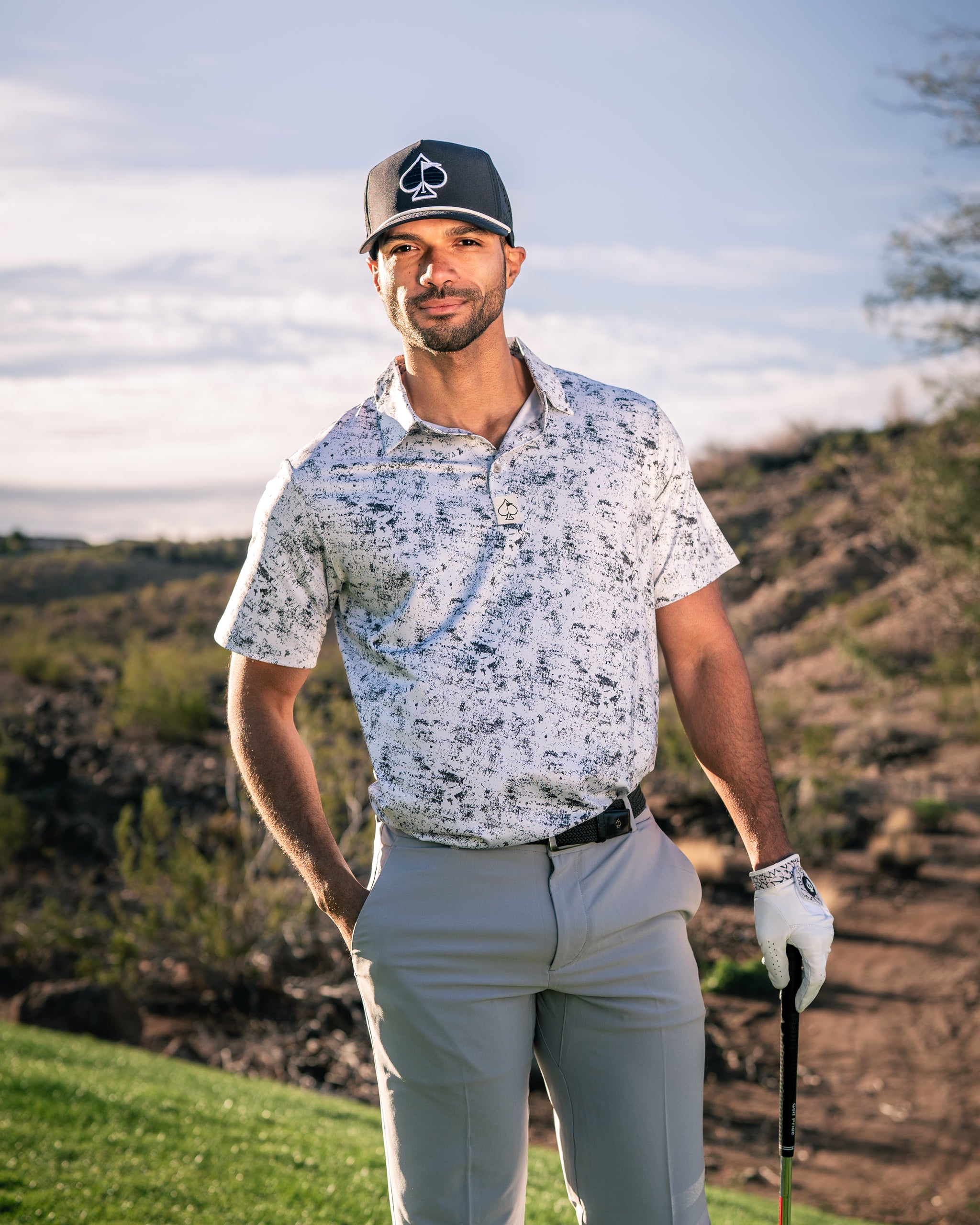 A golfer stands confidently on the course, wearing a stylish white noise-patterned polo shirt and gray pants, holding a golf club.