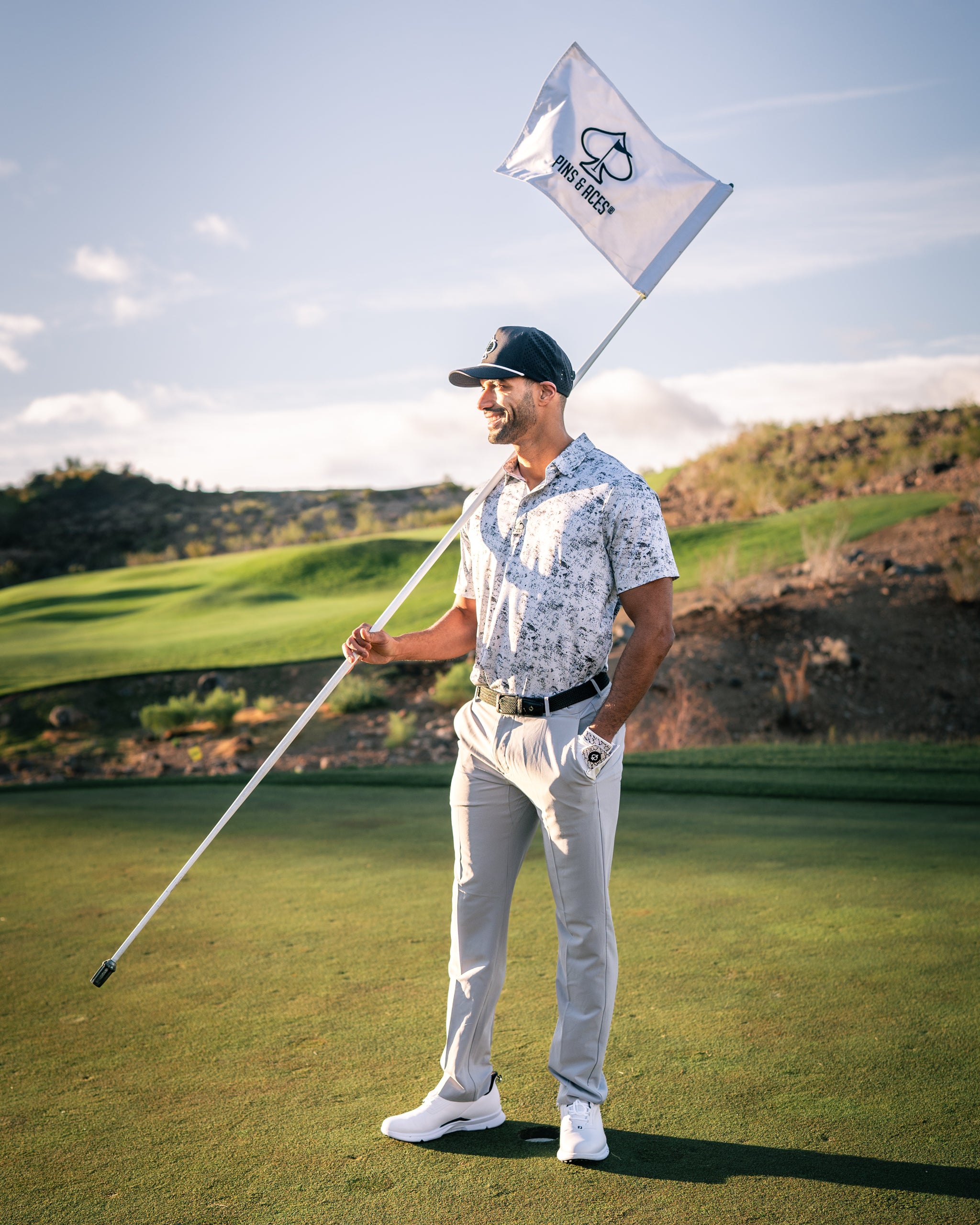 A golfer stands on a course holding a flag, wearing a stylish white patterned polo and light-colored pants.