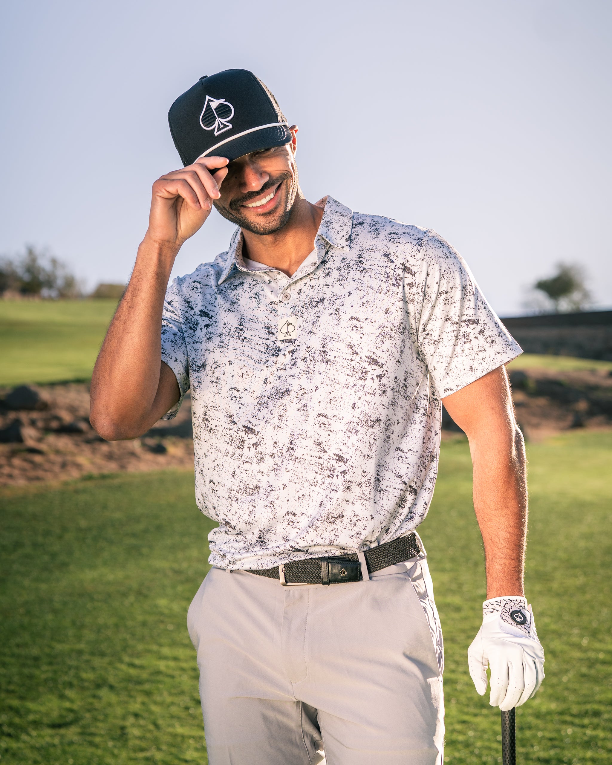 A golfer wearing a stylish white and gray patterned polo shirt and a black cap, smiling while holding a golf club on the course.
