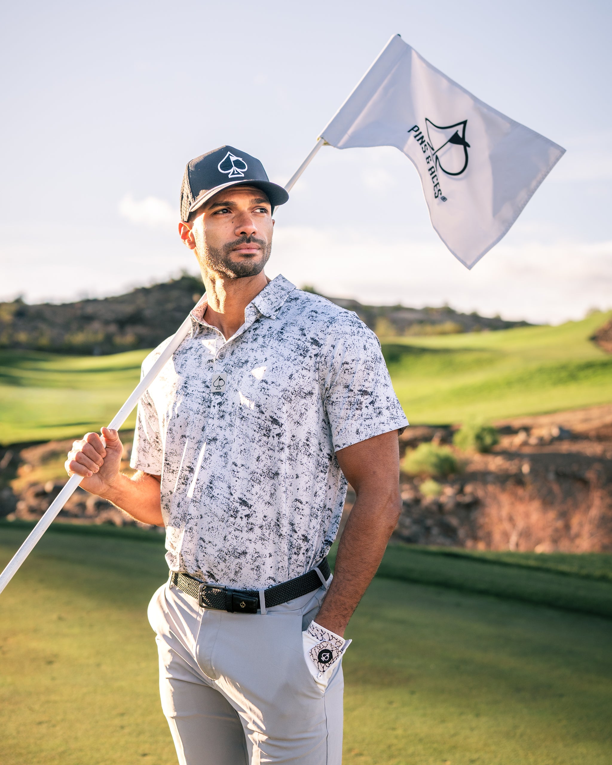 A golfer stands on the course holding a flag, wearing a stylish white and gray patterned polo shirt and a black cap.