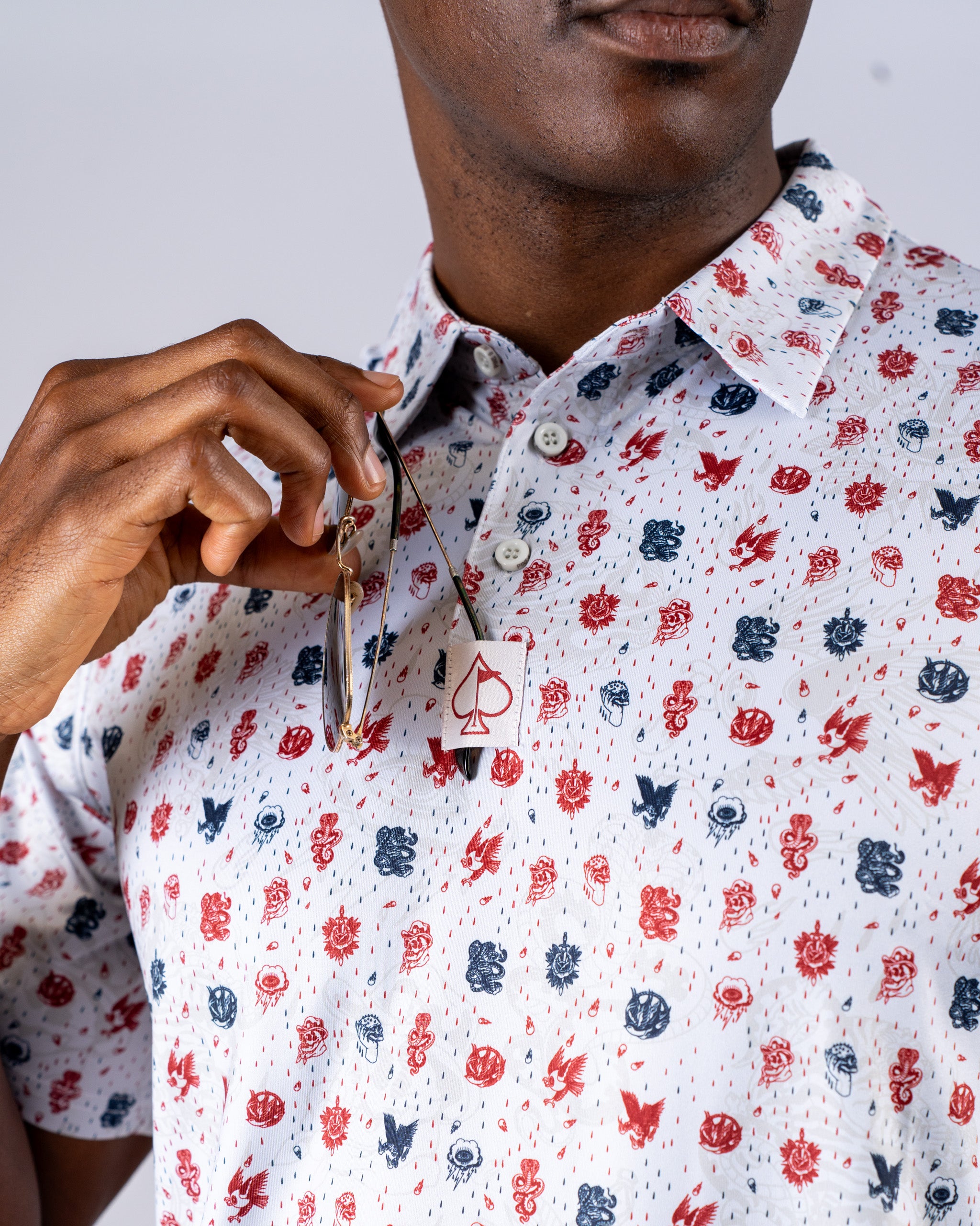 Model wearing an American Traditional golf shirt featuring a colorful pattern of iconic symbols, holding sunglasses.