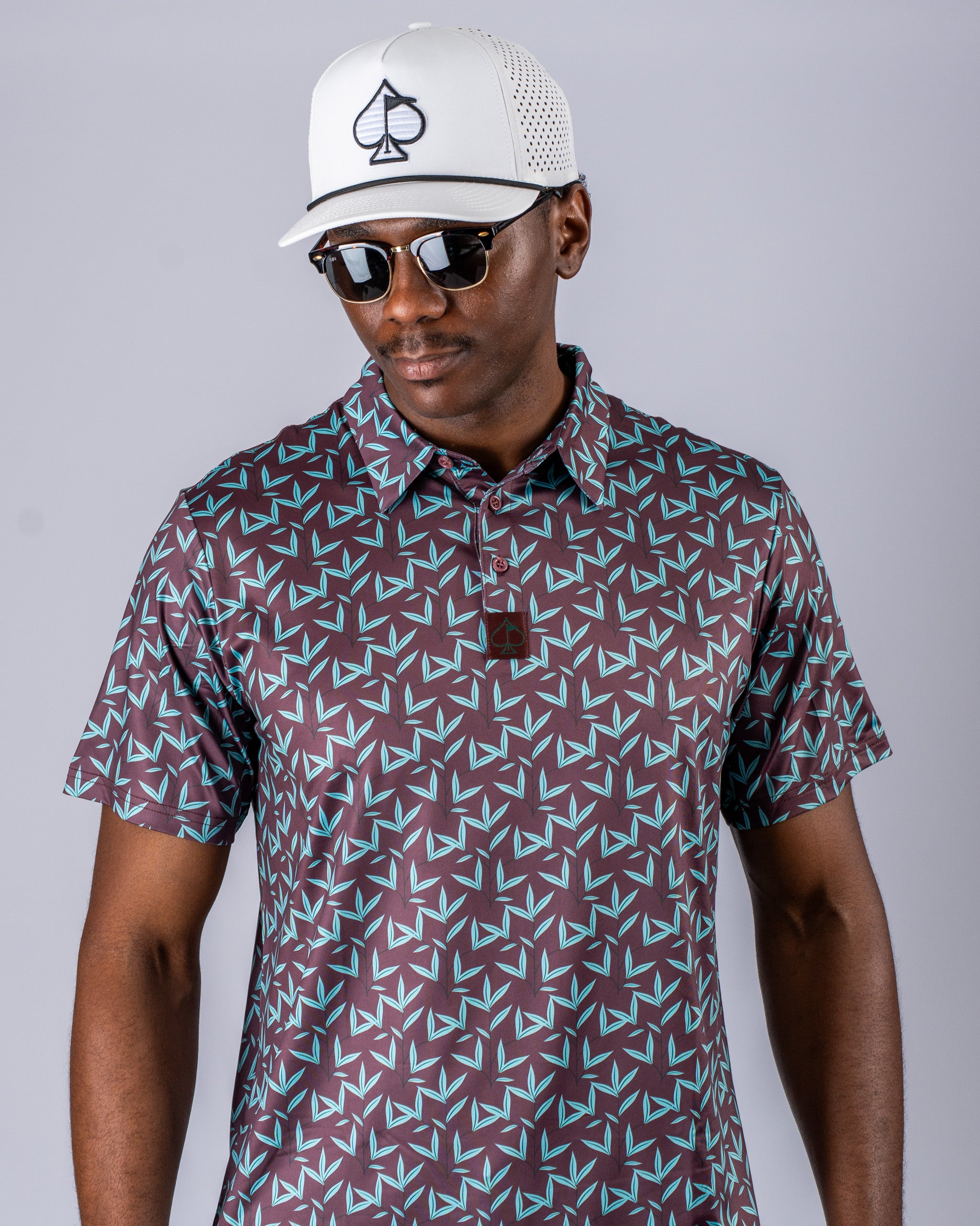 Men's Cocoa Verde golf polo featuring a bold geometric pattern in dark tones, paired with a white cap and sunglasses.