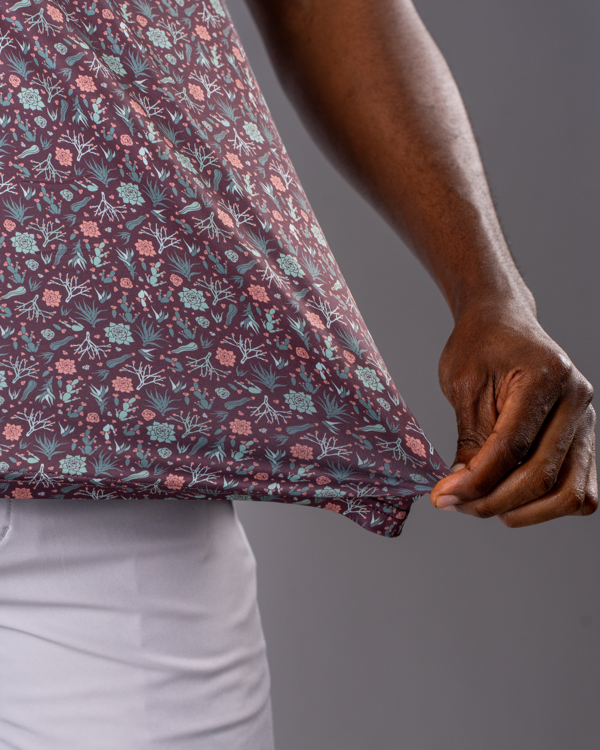 Close-up of a hand gripping the hem of a Desert Sage patterned golf shirt featuring floral designs in muted colors.