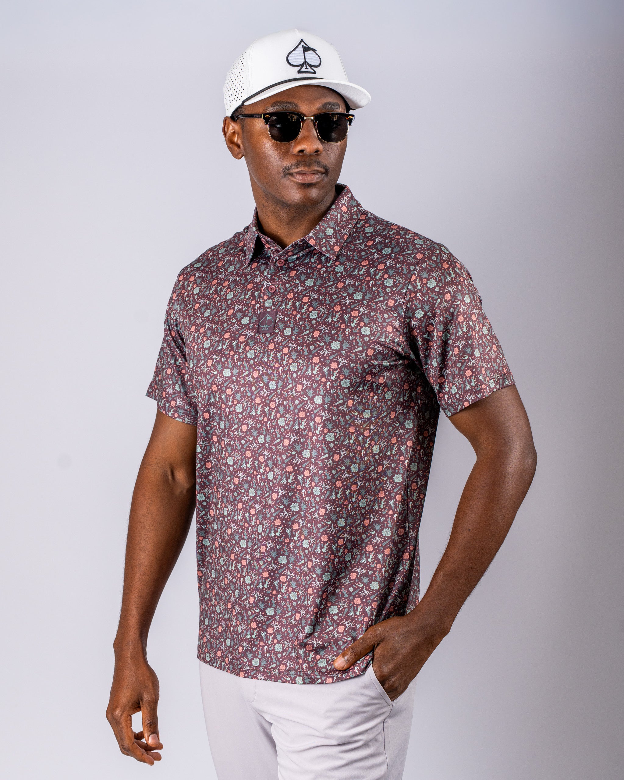 Men's short-sleeve golf shirt in Desert Sage pattern, paired with a white cap and sunglasses, showcasing a stylish look.