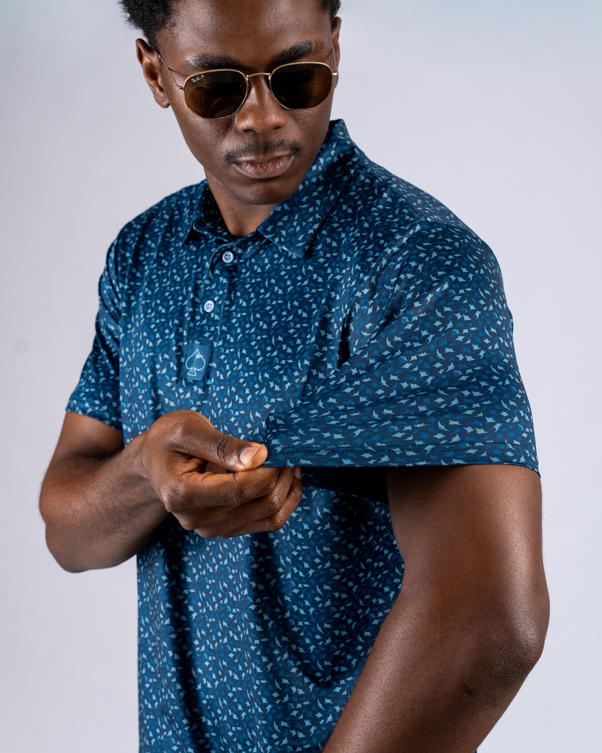Men's Manta Ray golf polo featuring a blue pattern, model adjusting sleeve while wearing sunglasses. Stylish and bold design.