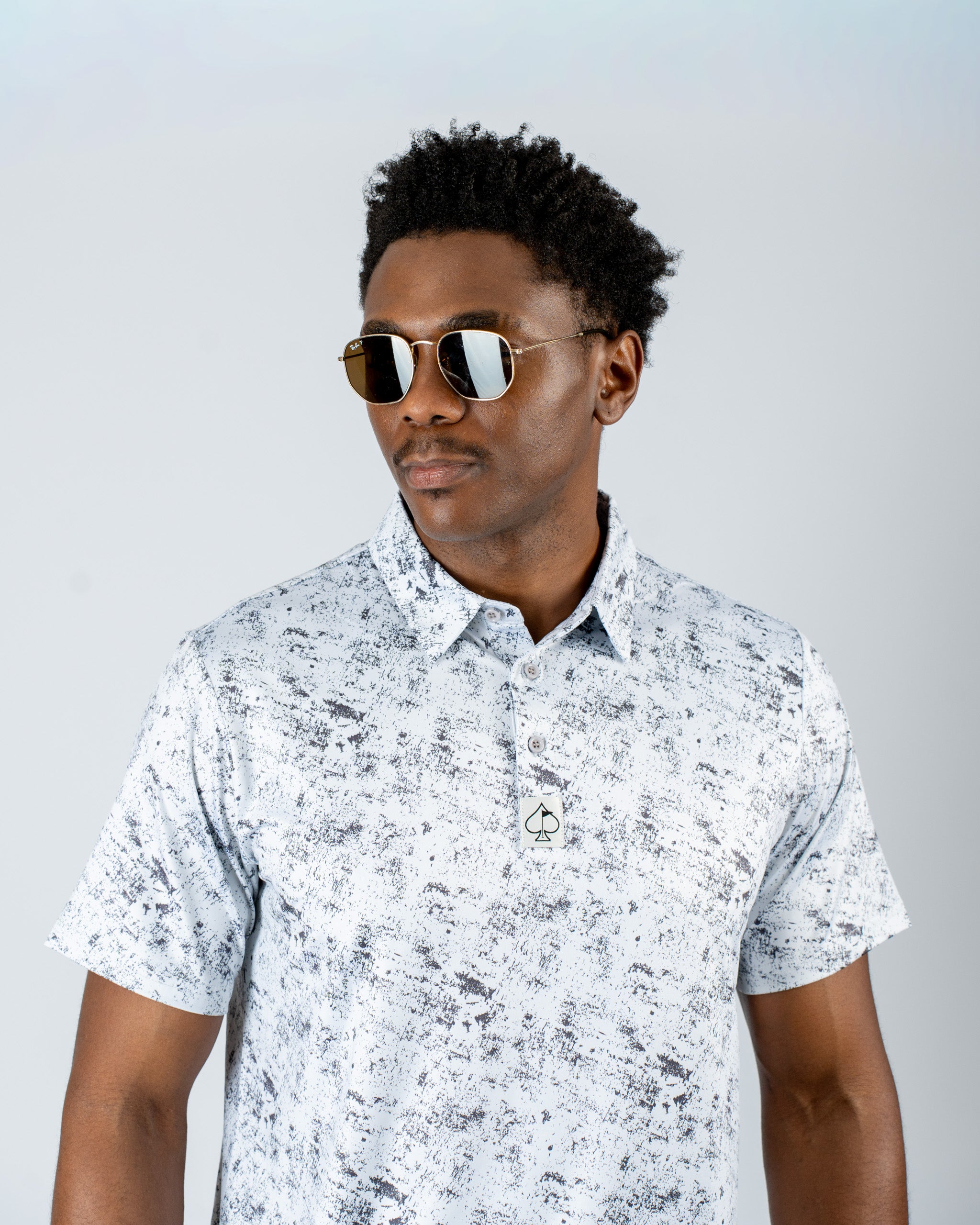 A man wearing a white and gray patterned golf polo shirt and sunglasses, showcasing a stylish and modern golf look.
