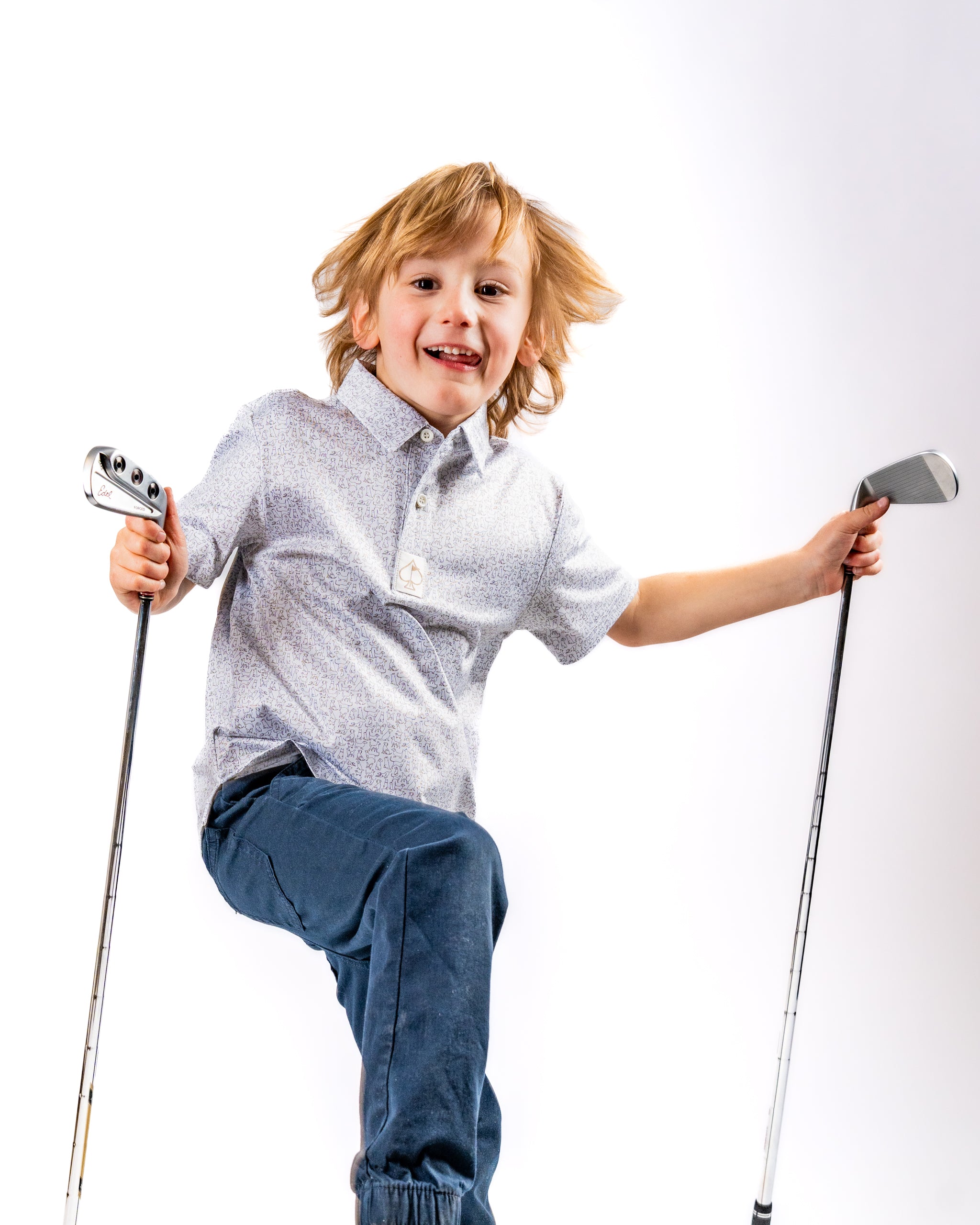 A joyful child in a patterned golf shirt poses with two golf clubs, showcasing a playful spirit on the course.
