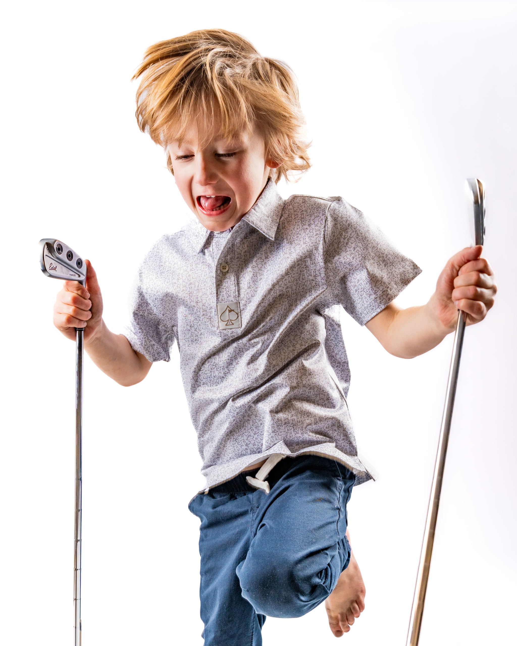 A young boy joyfully jumps while holding golf clubs, wearing a stylish light-colored shirt and casual pants.
