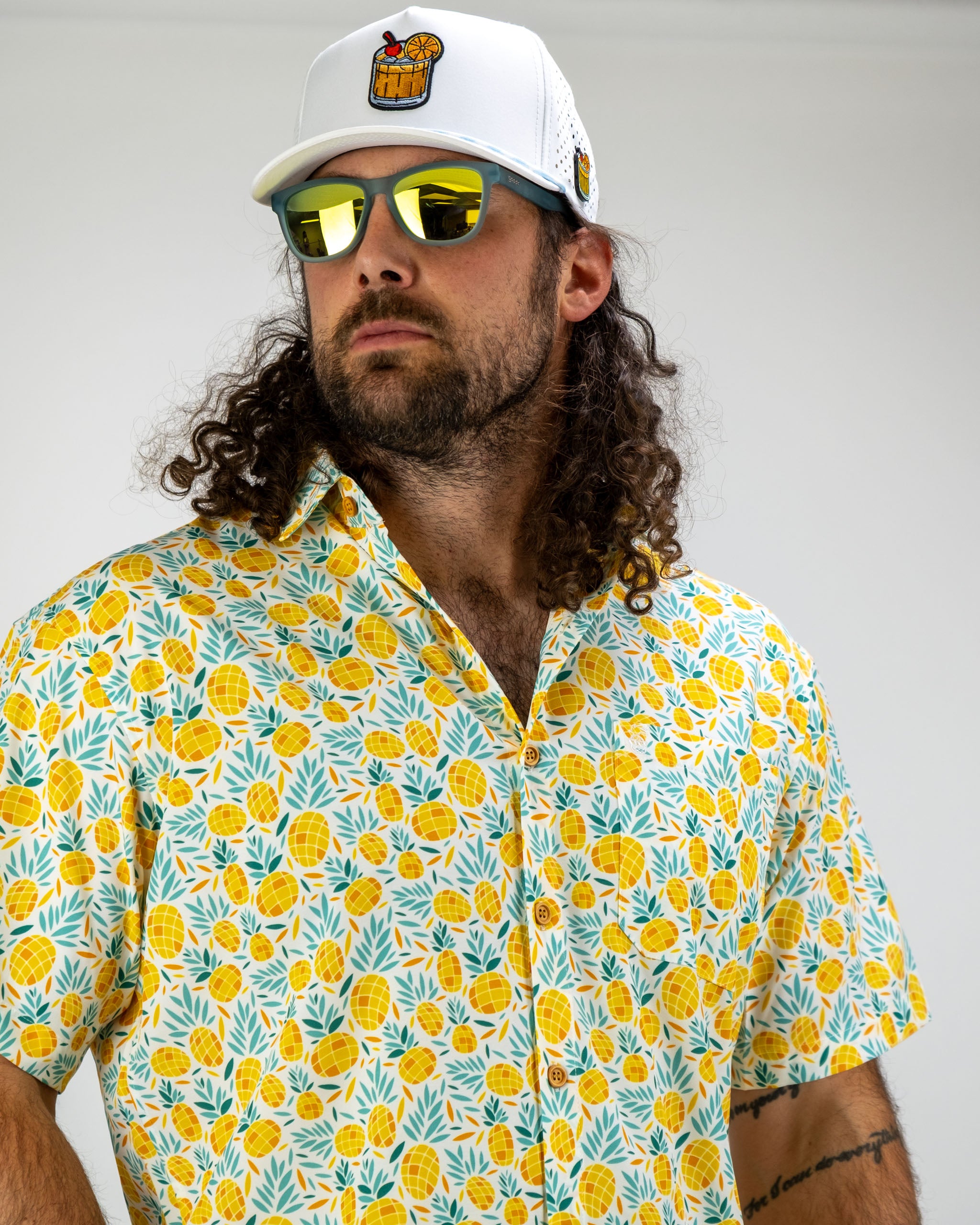 Man wearing a vibrant pineapple-patterned shirt and a white cap with a logo, showcasing a stylish golf look.