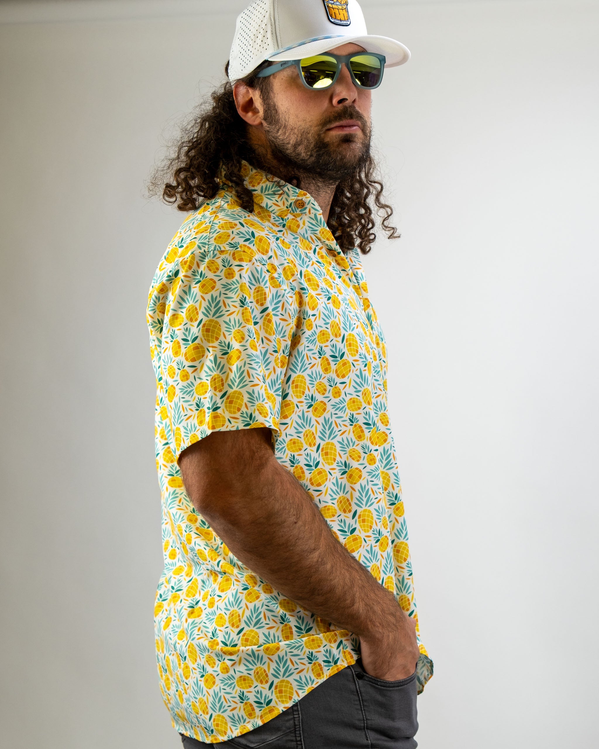 Man wearing a colorful pineapple-patterned shirt, sunglasses, and a white cap, posing against a neutral background.