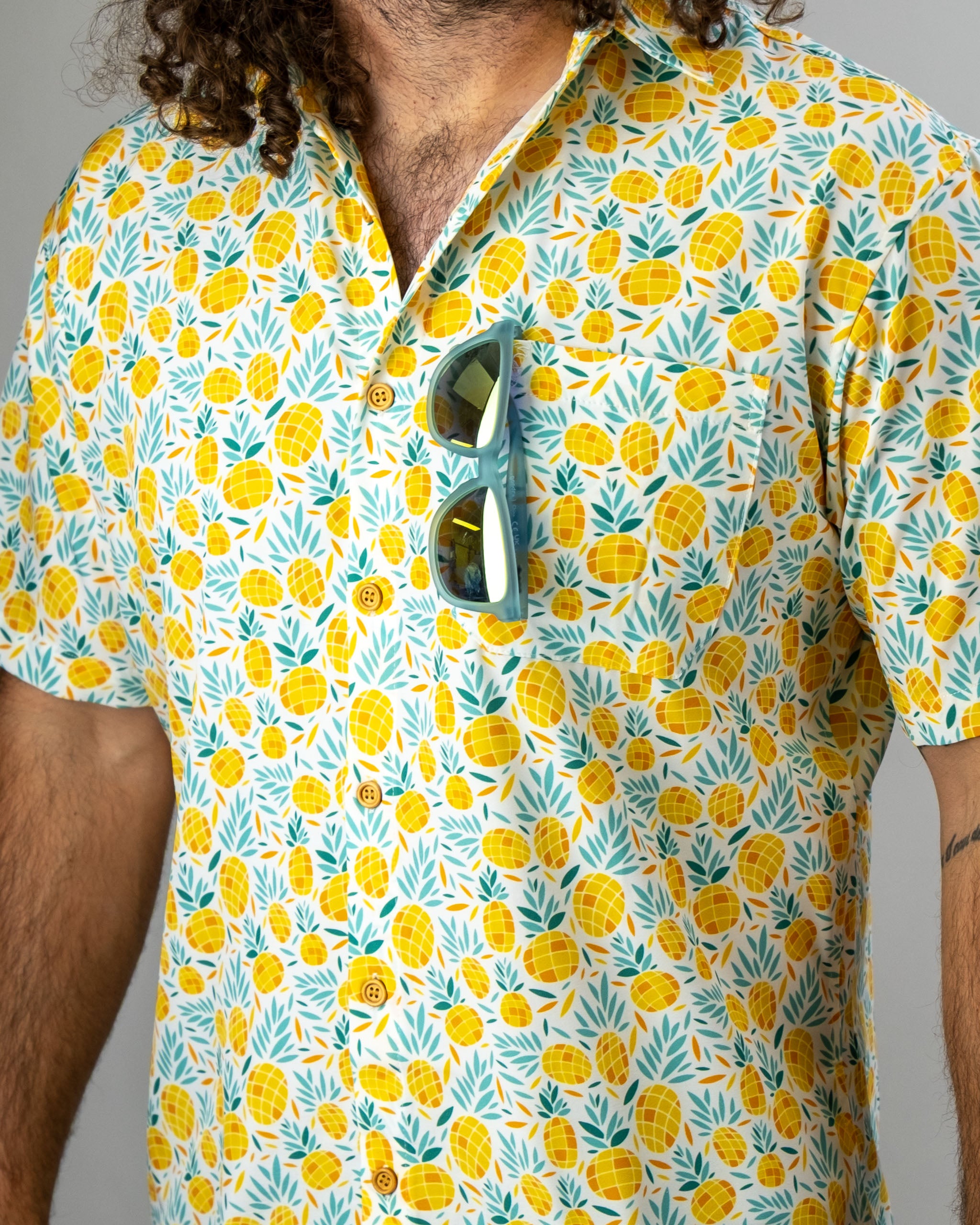 Colorful short-sleeve shirt featuring a pineapple pattern, worn by a person with curly hair and sunglasses tucked in.