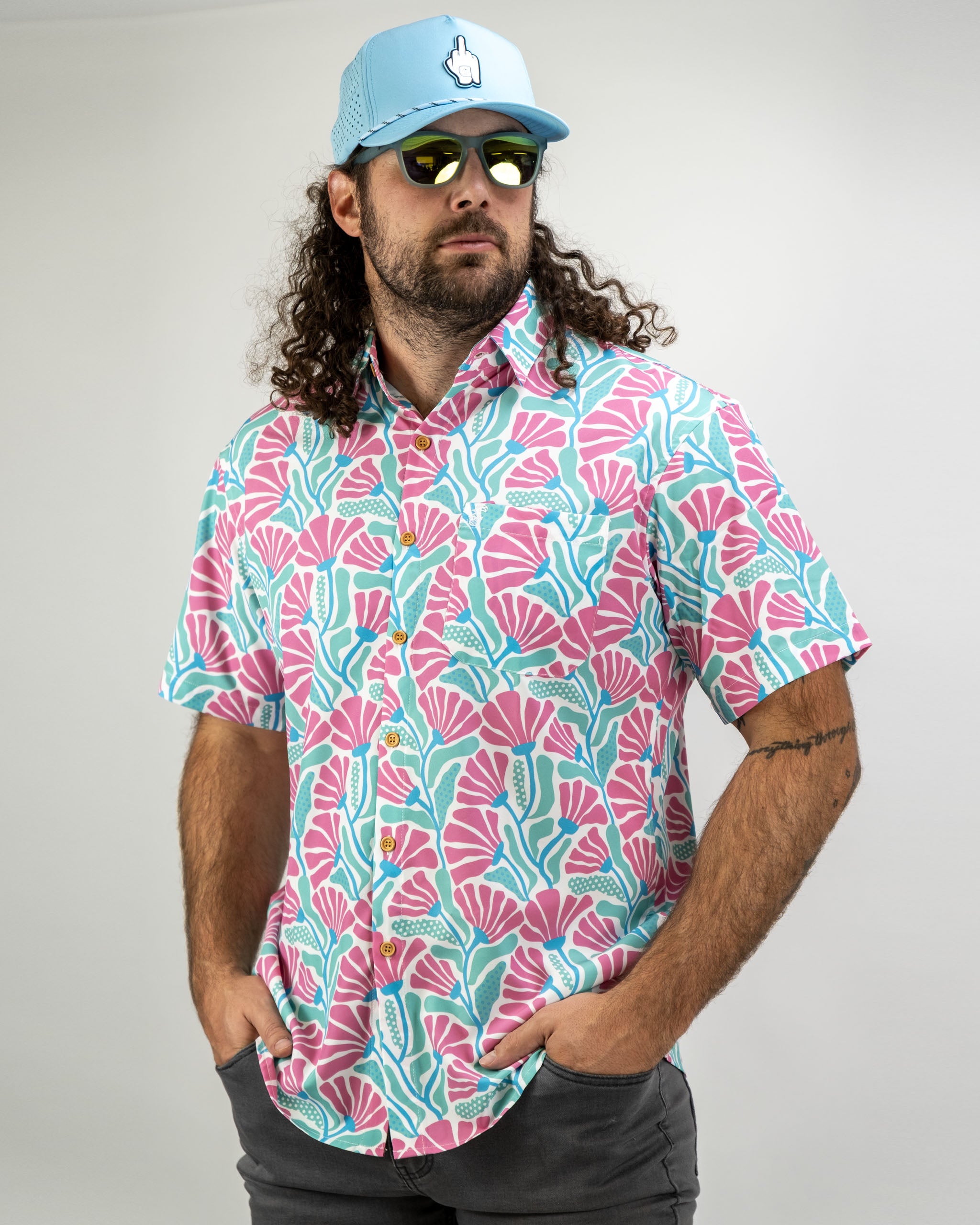 Bold coral and aqua patterned short-sleeve shirt worn by a man with curly hair, paired with a light blue cap and sunglasses.