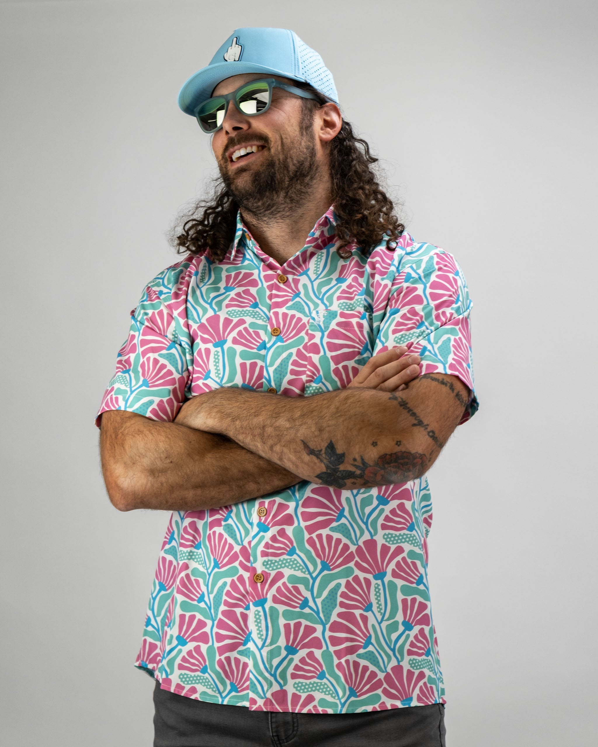 Bold coral and teal floral shirt worn by a man with curly hair, paired with a light blue cap and sunglasses.