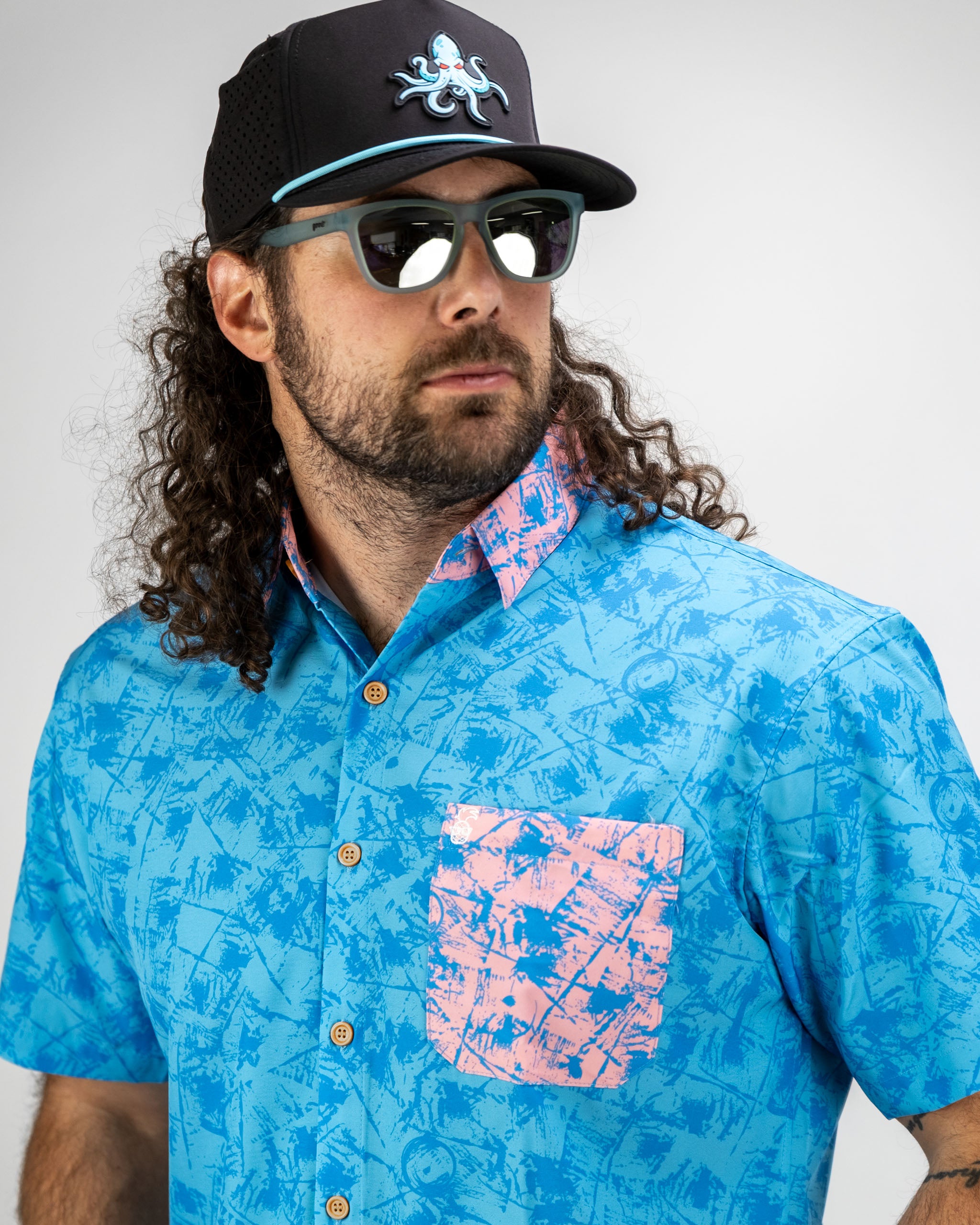 A man wearing a blue patterned short-sleeve shirt with a pink pocket, paired with a black cap and sunglasses.