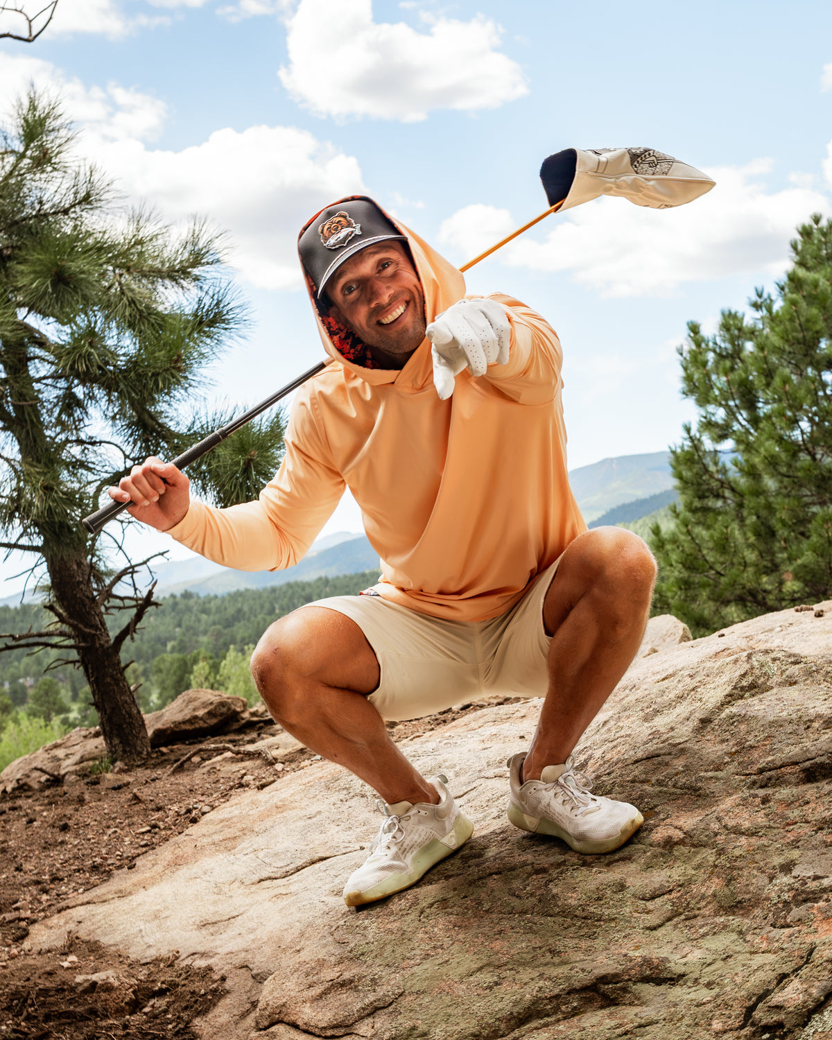 Man wearing an autumn leaves performance golf hoodie, posing with a golf club on a rocky outdoor setting.