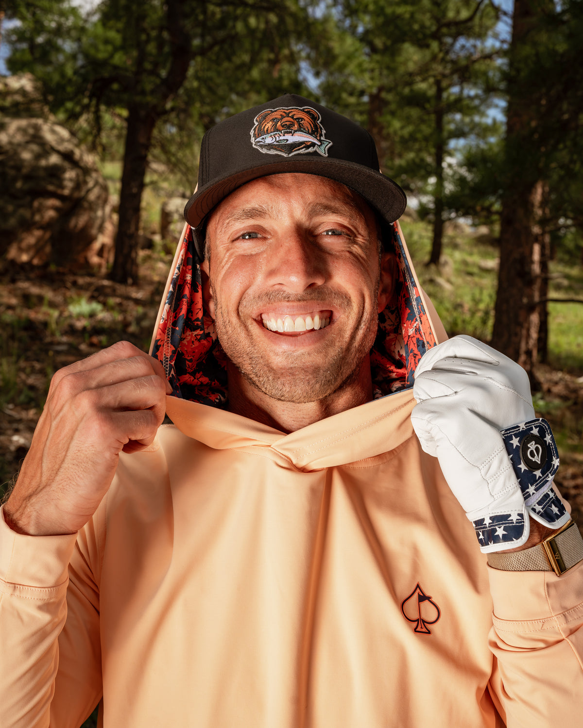 Man wearing a peach performance golf hoodie with a vibrant autumn leaves lining, smiling outdoors in a wooded area.