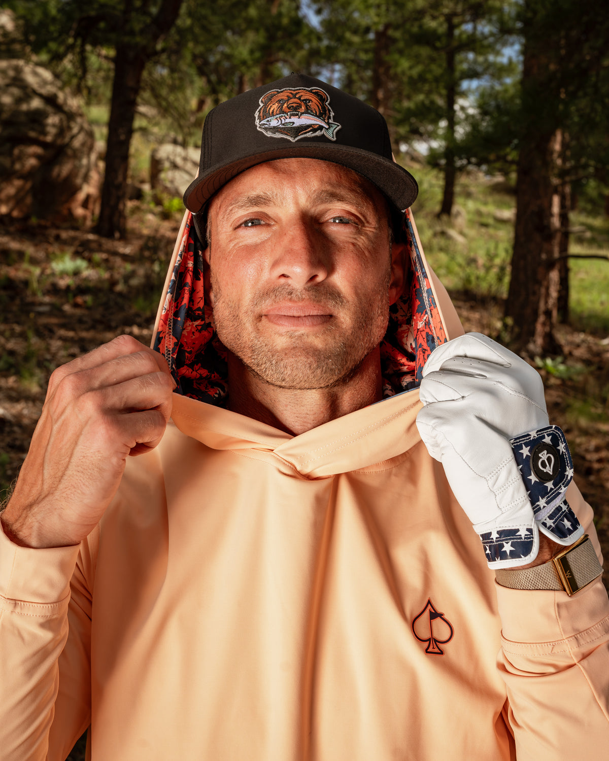 Man wearing a peach performance golf hoodie with a floral-lined hood, holding a golf glove and cap in a natural setting.