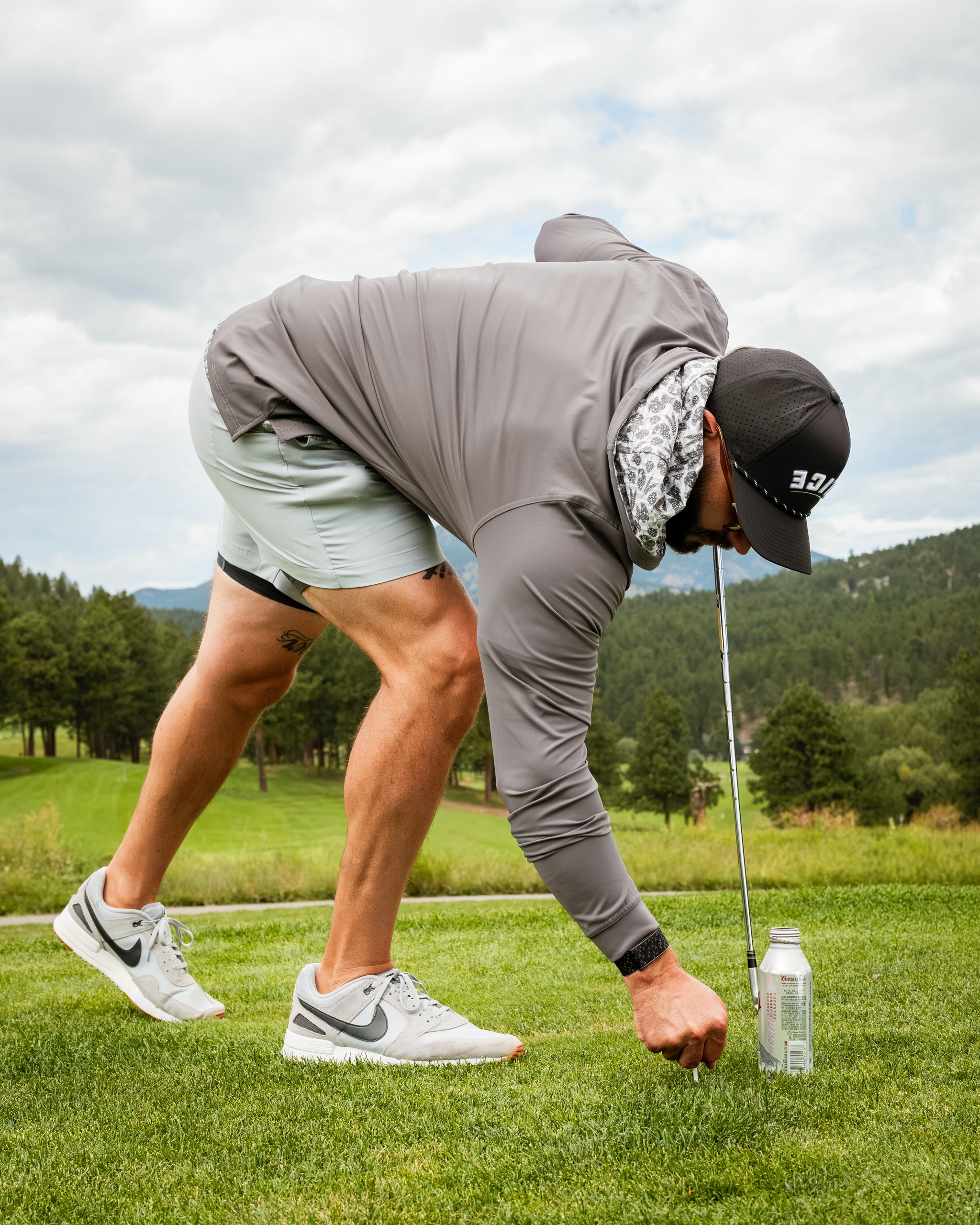 Man in a performance golf hoodie bending down on a golf course, preparing to tee off with a can nearby.