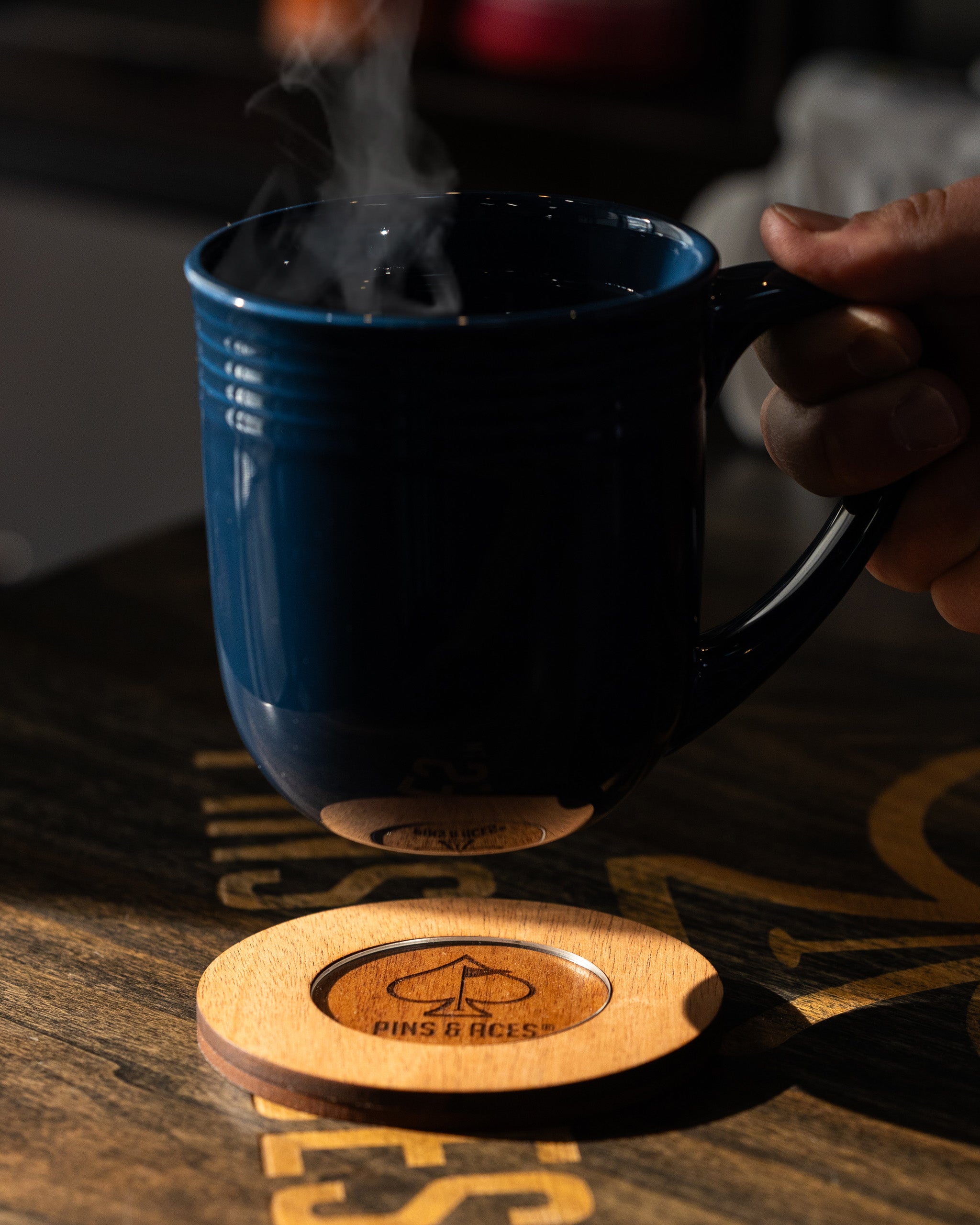 Dark blue ceramic mug with steam, held by hand, resting on a wooden coaster with "Pins & Aces" logo, for bold golf style.