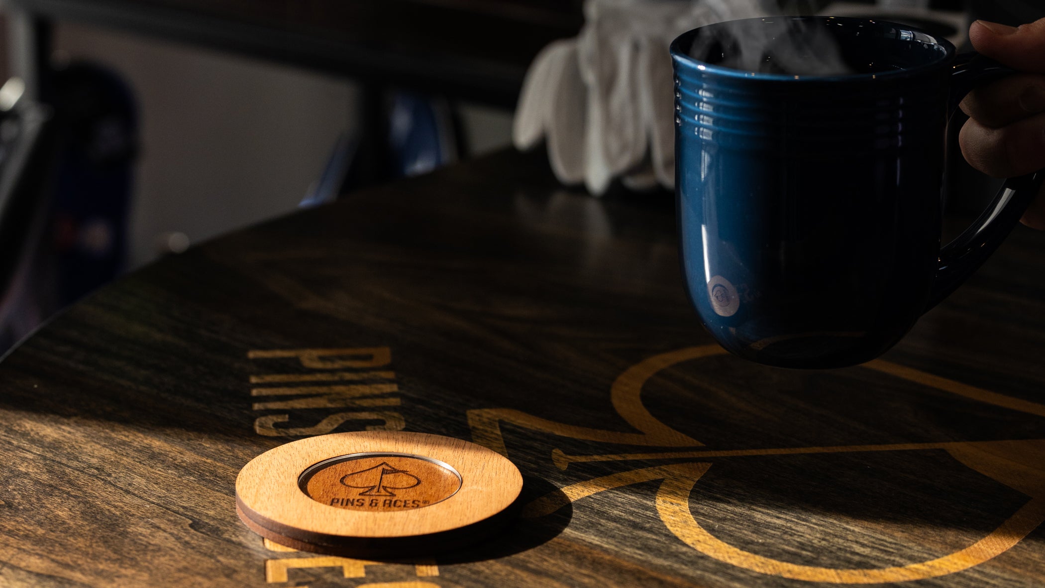 Blue ceramic mug held by hand, placed on a dark wooden table with "Pins & Aces" logo coaster nearby, highlighting bold gol...
