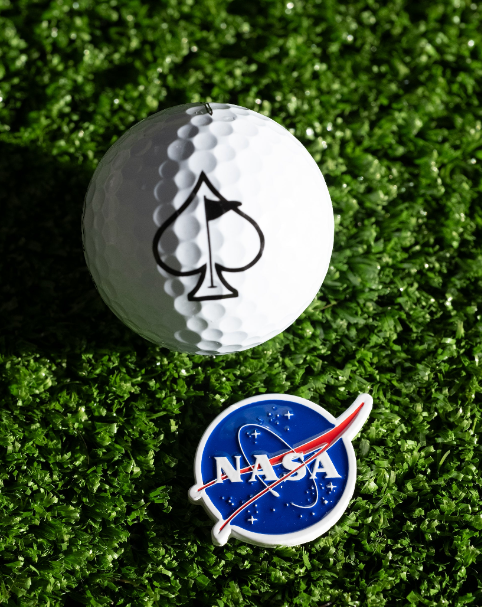 NASA "Meatball" ball marker featuring the iconic logo, placed next to a golf ball with a spade symbol on a green surface.