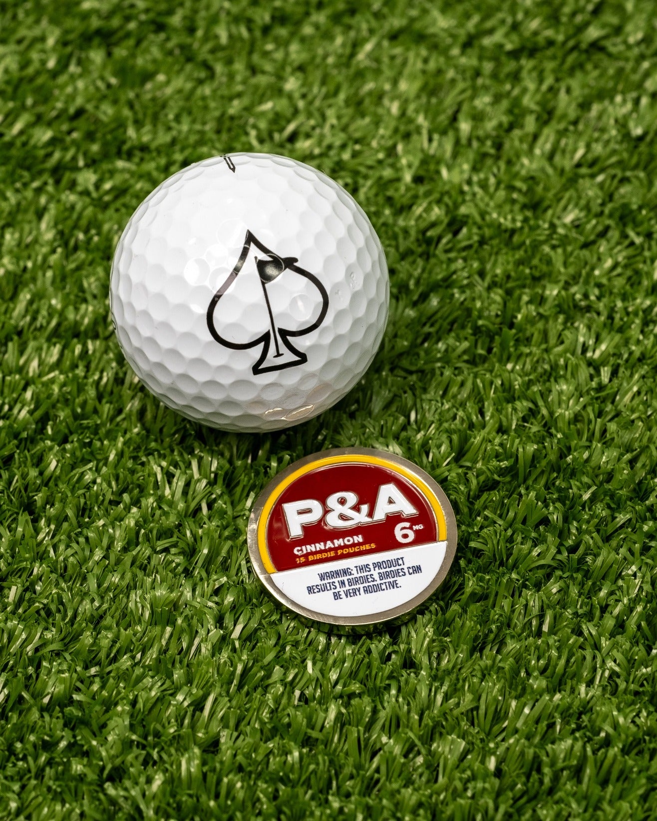 White golf ball with an ace of spades logo next to a round cinnamon ball marker featuring bold red and yellow design.