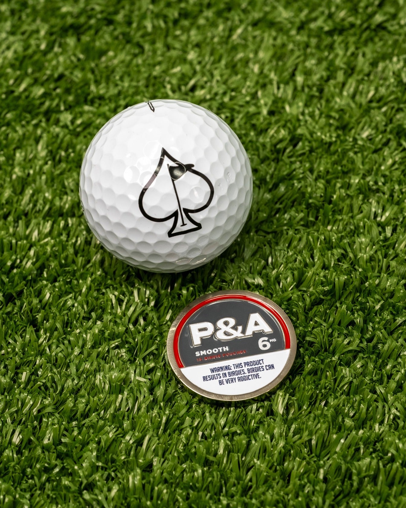Smooth ball marker with a bold design featuring the P&A logo, placed next to a white golf ball on green turf.