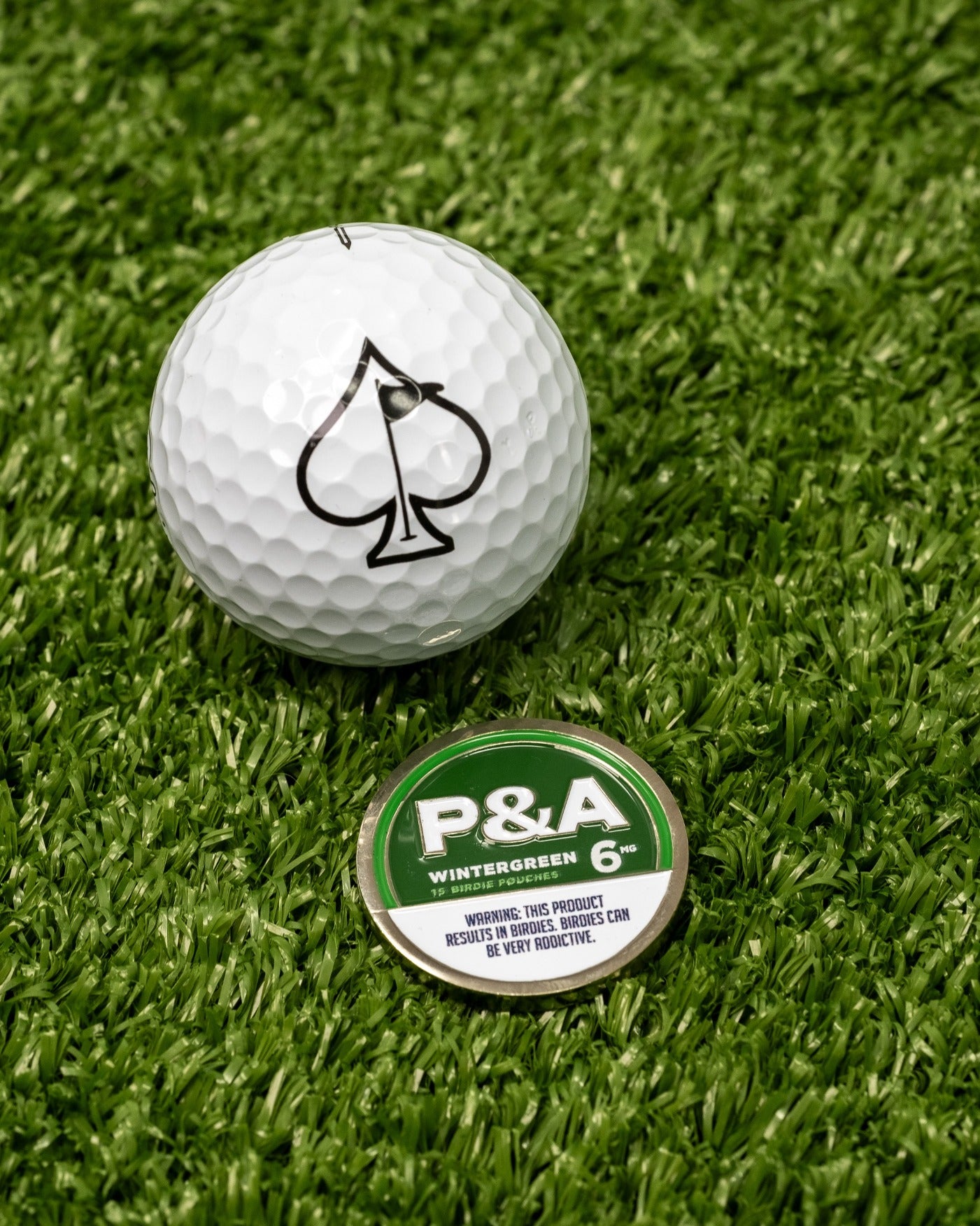 Wintergreen ball marker beside a golf ball featuring a spade logo, set on a textured green grass background.