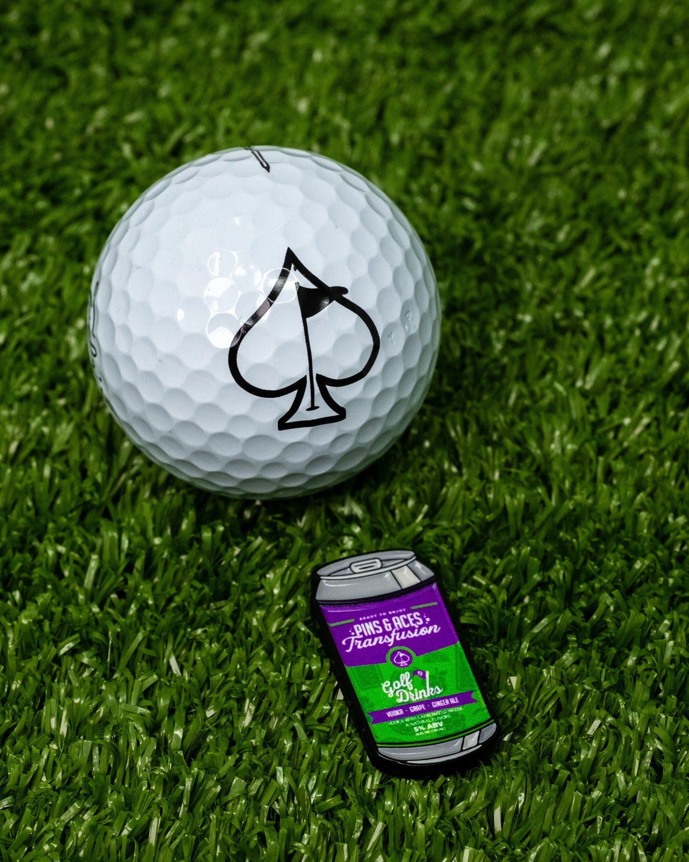 Transfusion Can Ball Marker next to a golf ball with a spade logo, set on green turf. Perfect for stylish golfers.