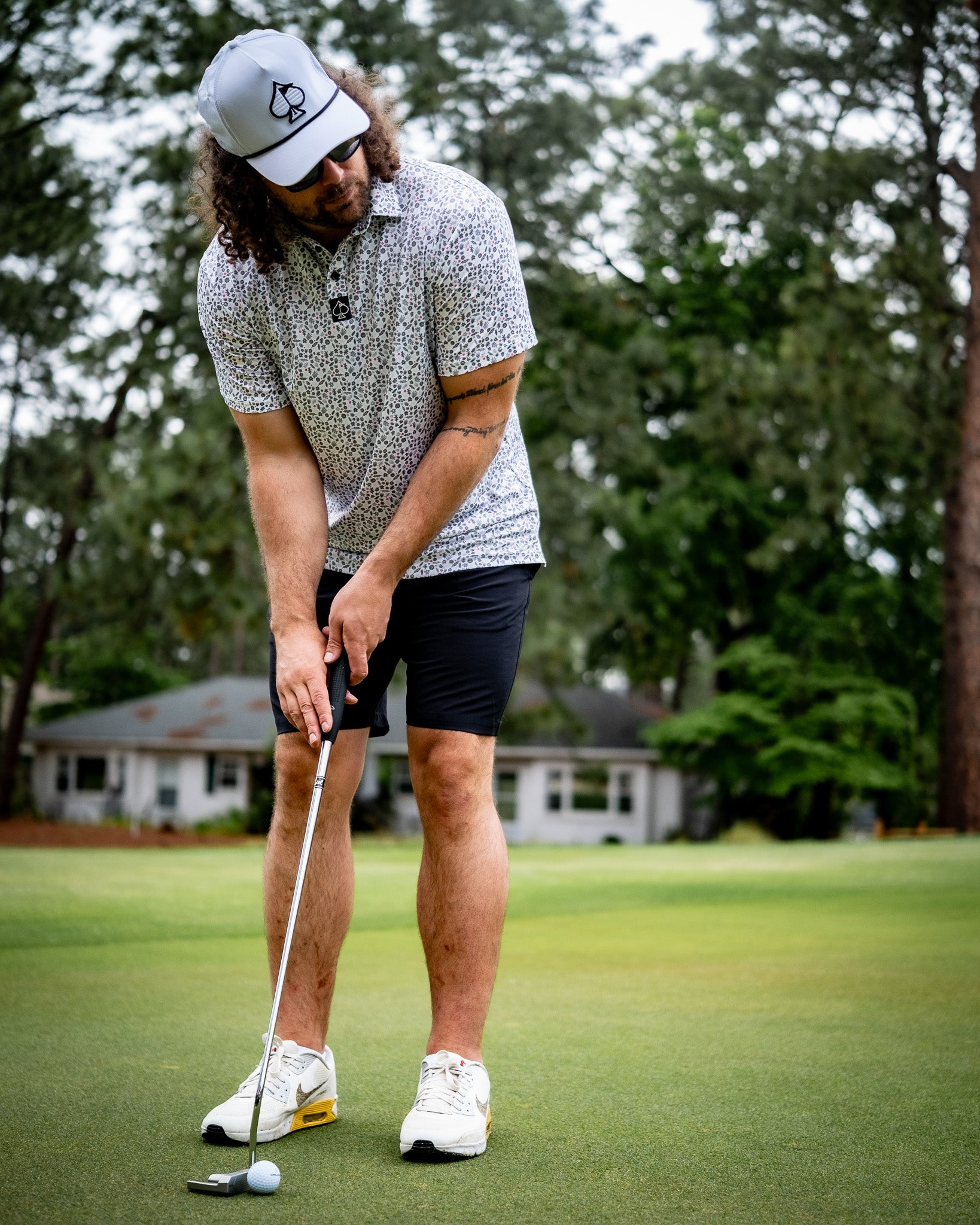 A golfer wearing black performance shorts and a patterned polo shirt, putting on a green golf course.