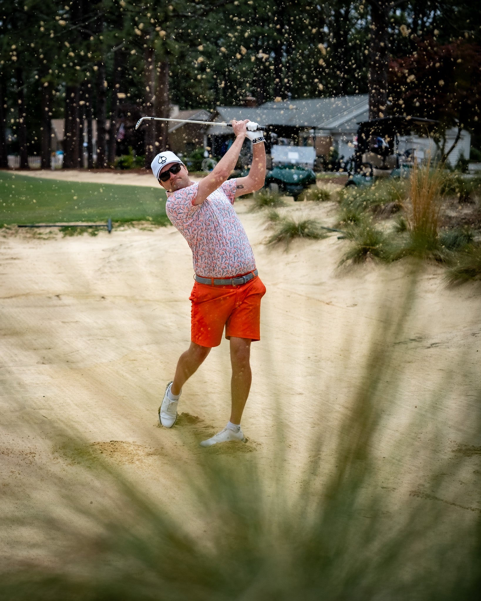 A golfer in melon-colored performance shorts swings a club from a sand trap, with sand flying around him.