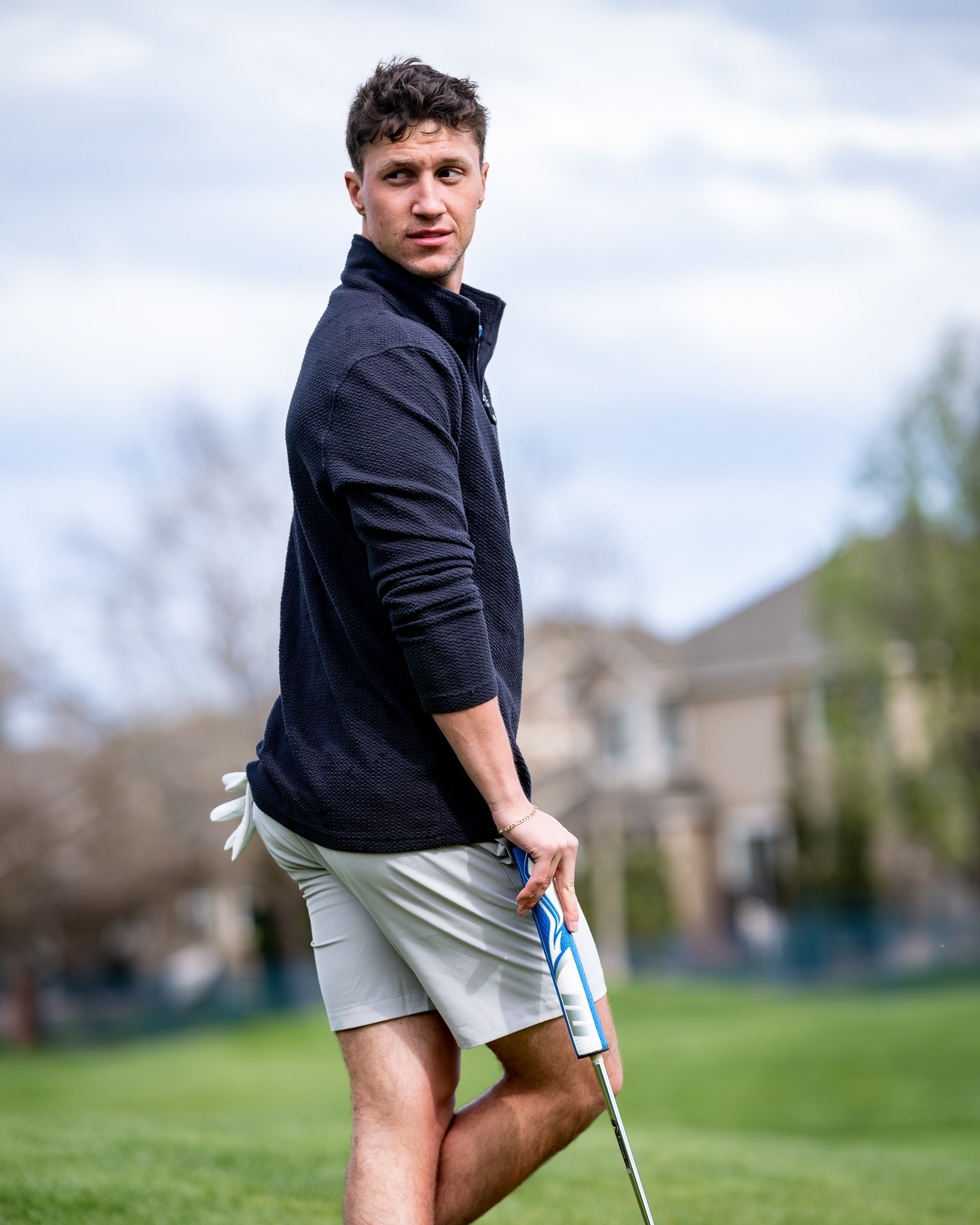 A golfer wearing dark stone performance shorts and a navy pullover stands on the course, holding a golf club.
